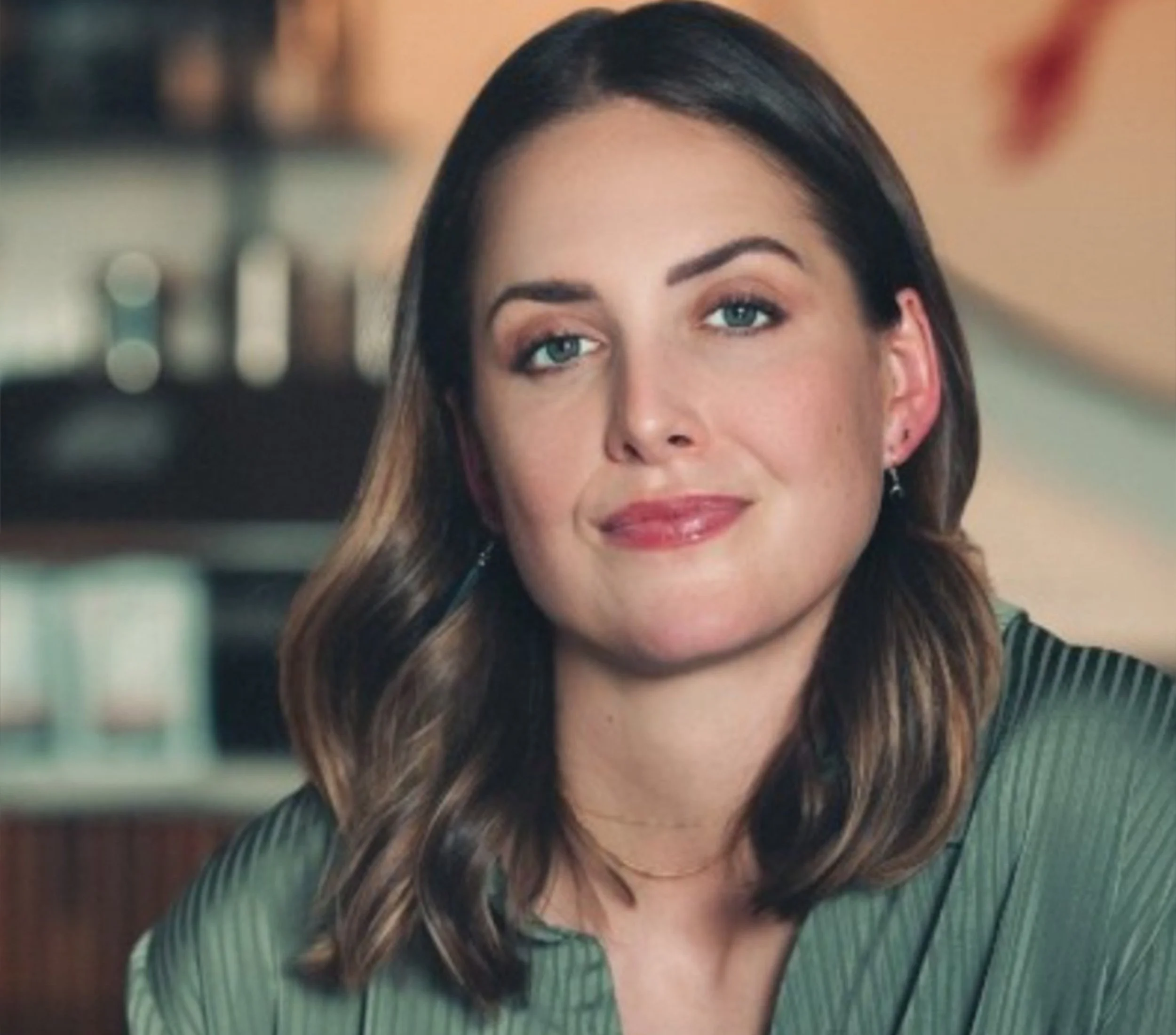 Close-up of a woman with long brown hair, light blue eyes, and light makeup, wearing a dark green striped top, in an indoor setting.