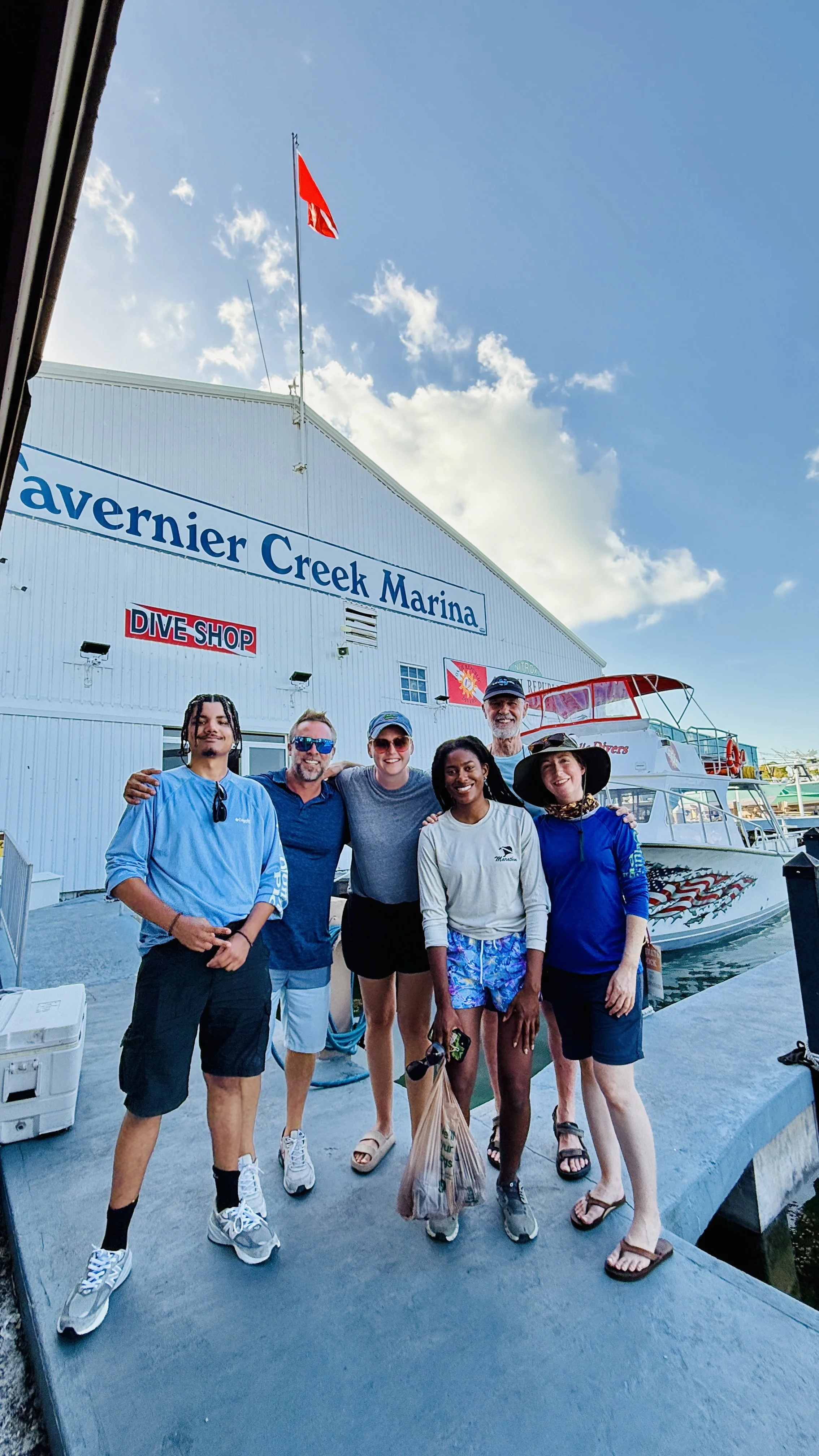 A group of six people standing together on a dock at the marina, smiling for the photo, with boats and the marina building in the background.
