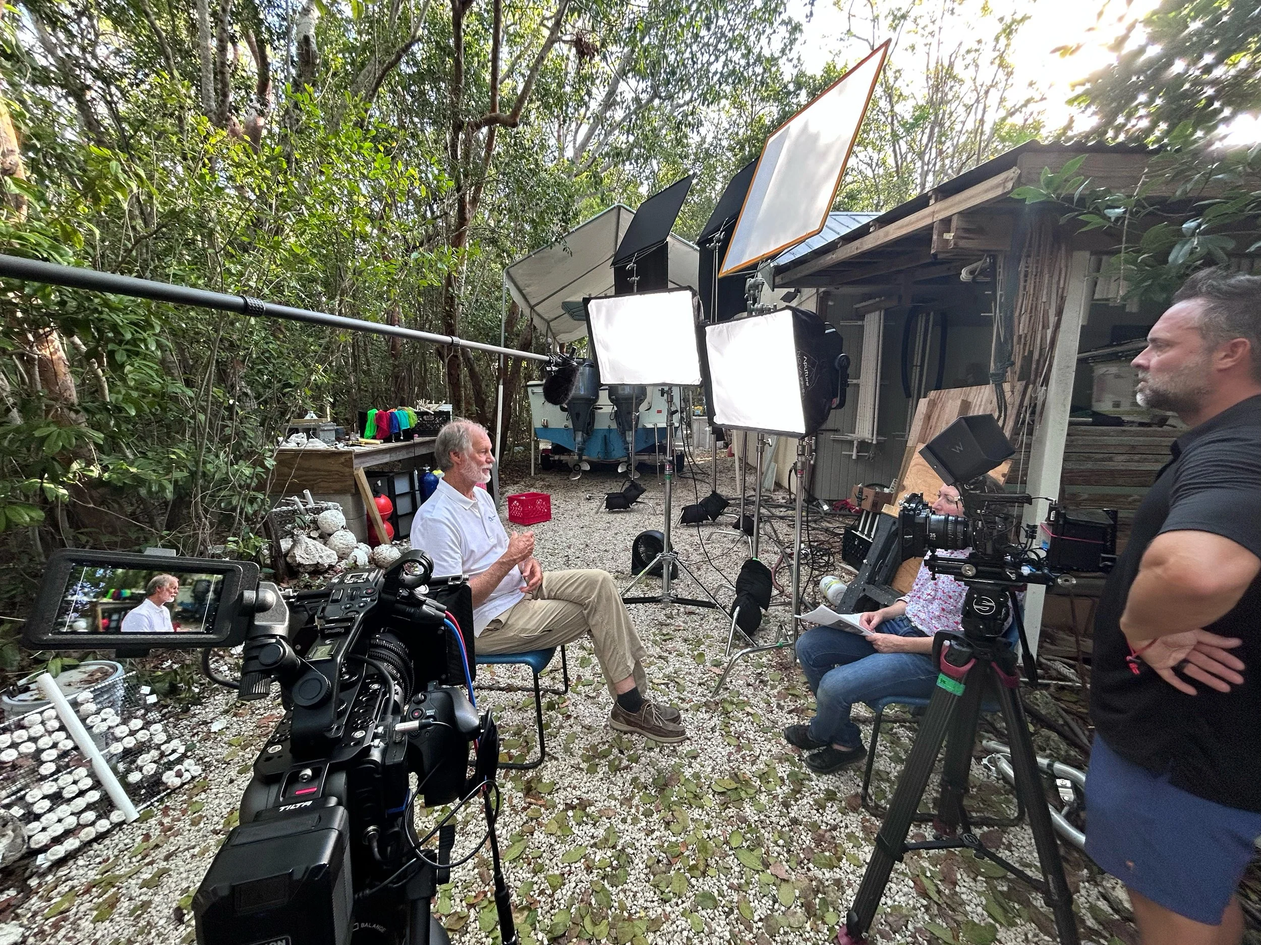A behind-the-scenes view of a film or video shoot in an outdoor wooded area with professional filming equipment, lights, and crew members, including an older man sitting and a woman holding a script, surrounded by trees and natural light.