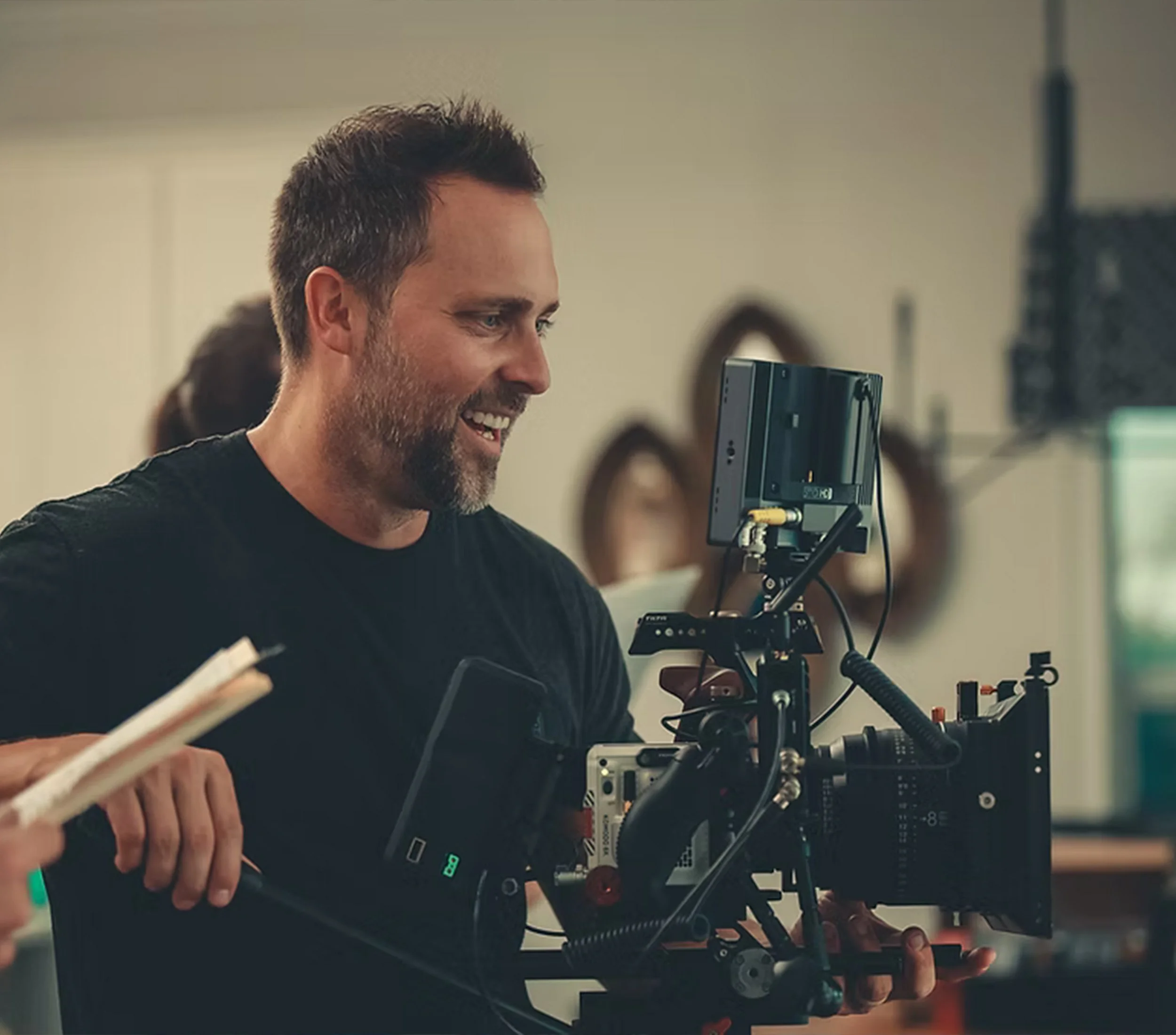 A man with dark hair and a beard watching a monitor with a smile in a film or television production setting, with a person holding a clapperboard in the foreground.