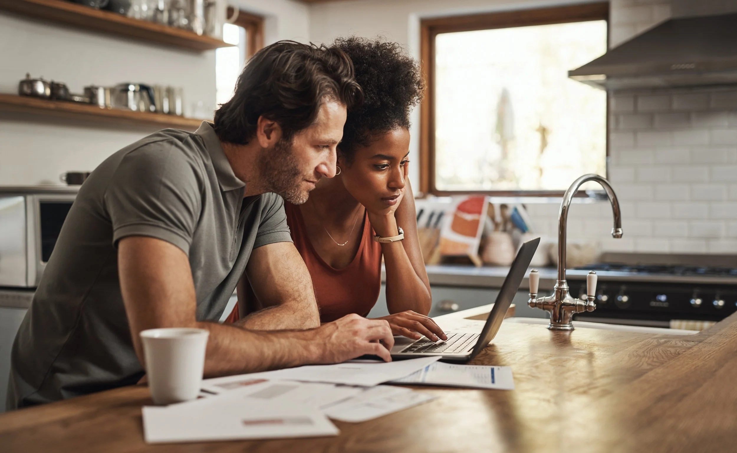 Couple using a laptop in a kitchen with papers and a cup on the countertop.