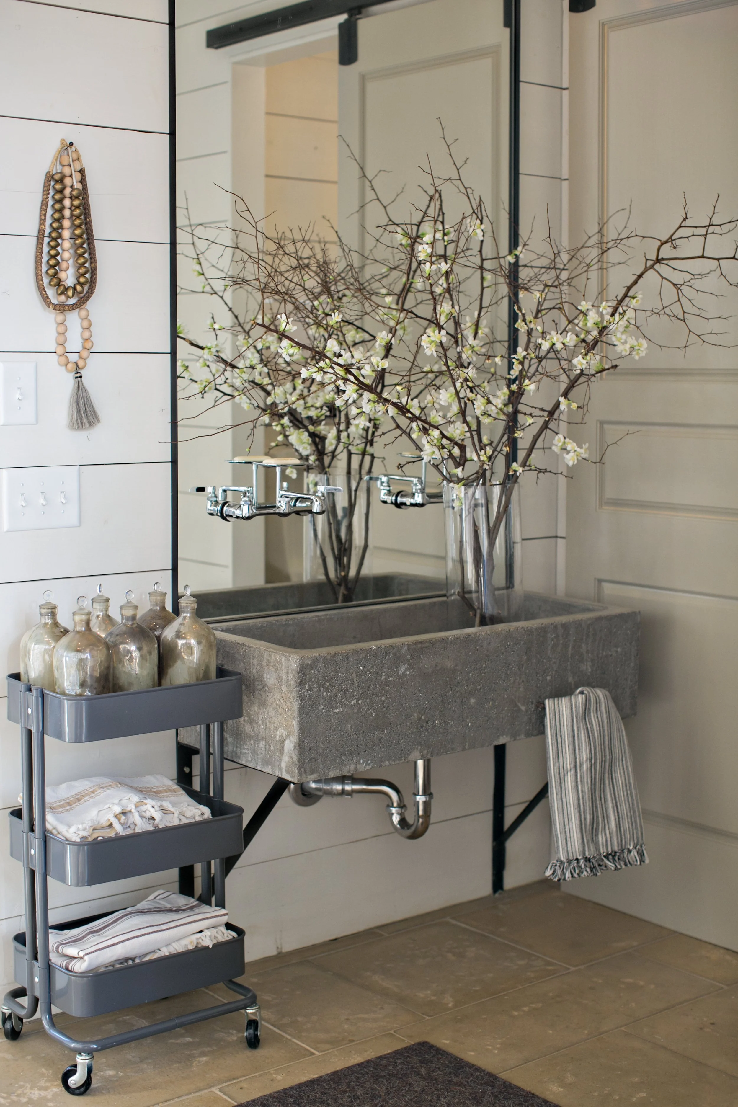 Decorative bathroom sink with a concrete basin, large mirror, and flower arrangements in vases, with a cart holding glass bottles and towels in a home interior.