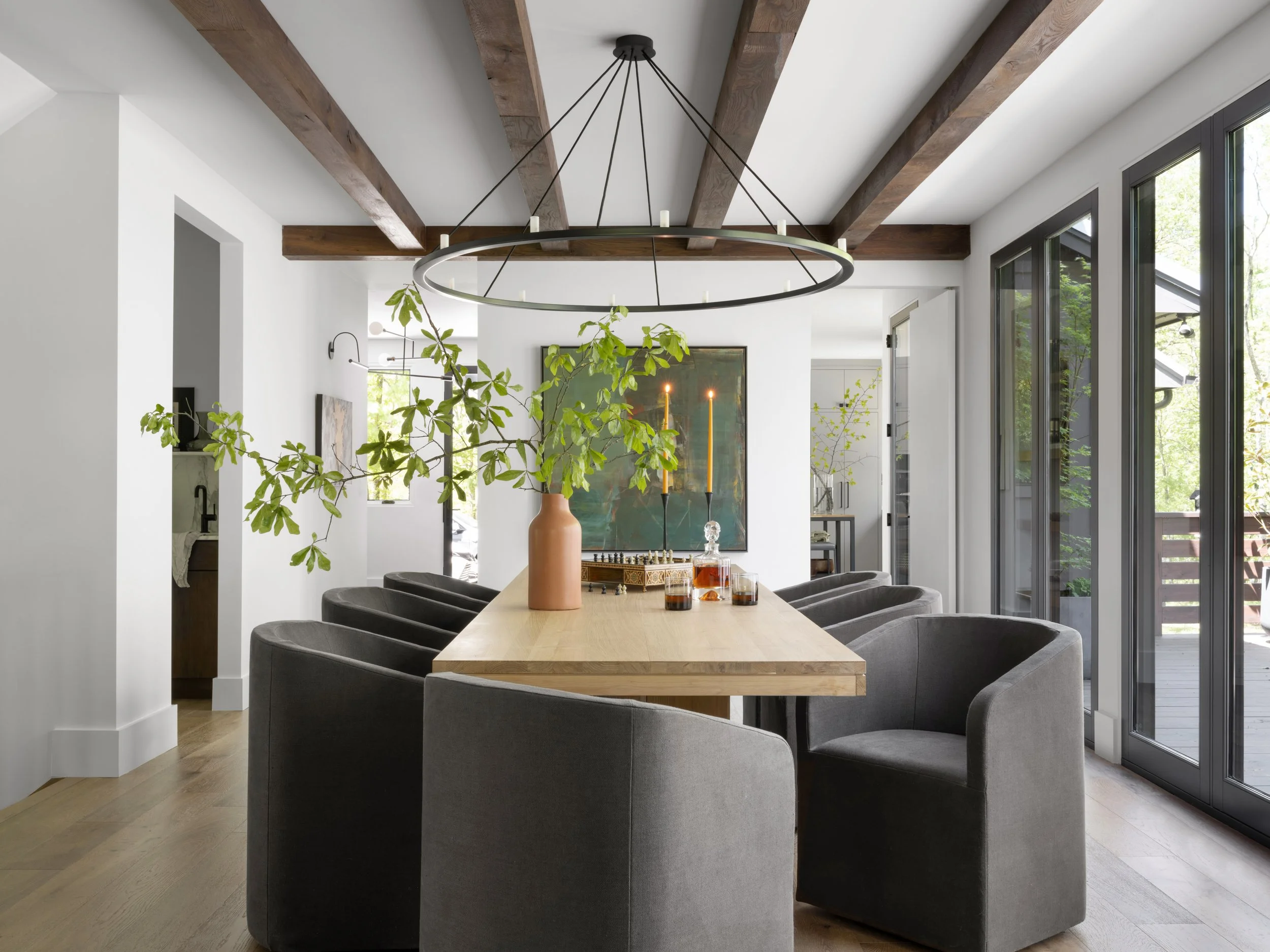 Modern dining room with a wooden table, eight gray upholstered chairs, a large circular chandelier, and natural light streaming in through large windows and glass doors. Decor includes a vase with green foliage, candles, and artwork on the walls.