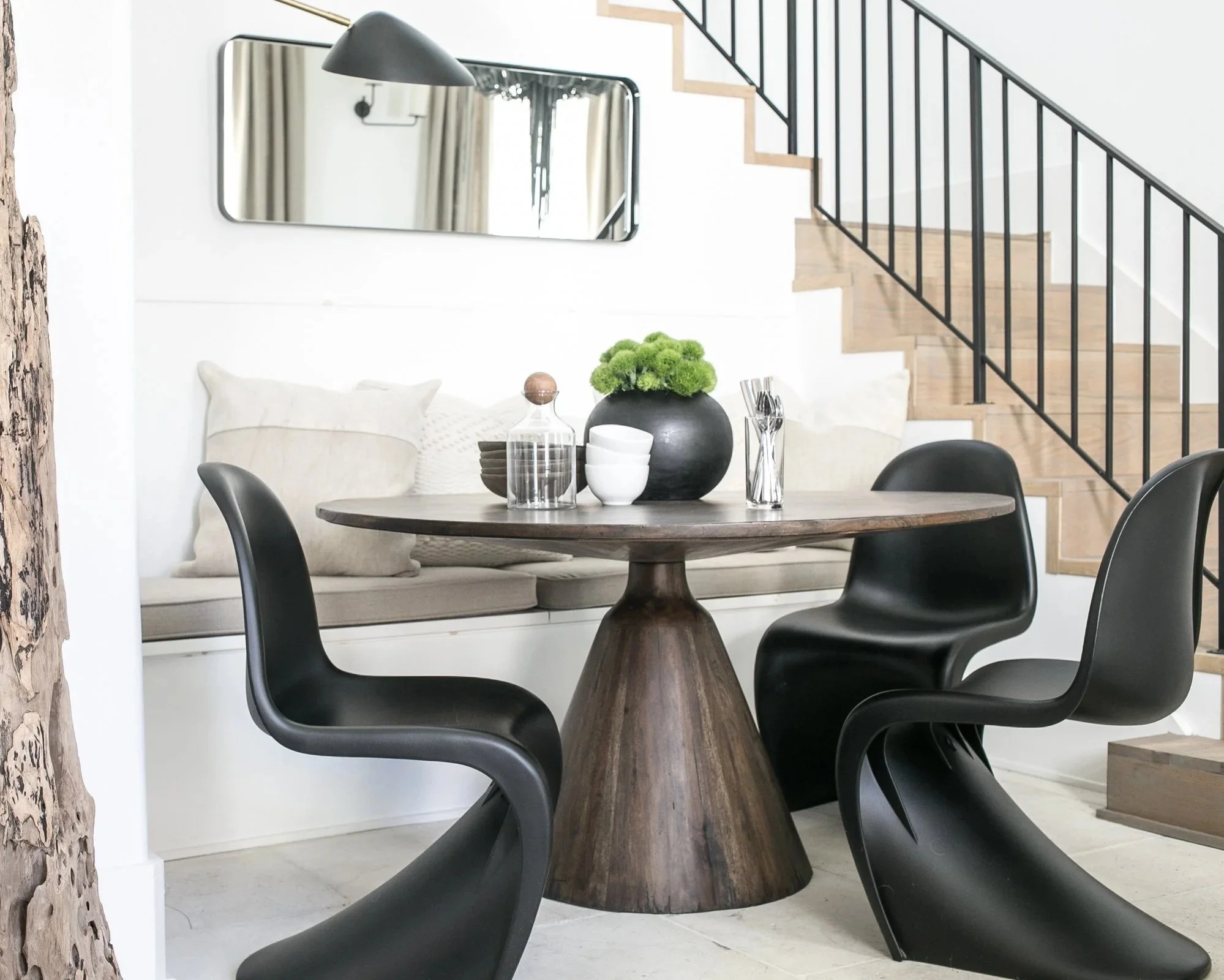Modern dining area with a round wooden table, three black molded plastic chairs, a white built-in bench with cushions, a black vase with green flowers on the table, and a staircase with wooden steps and black metal railing.