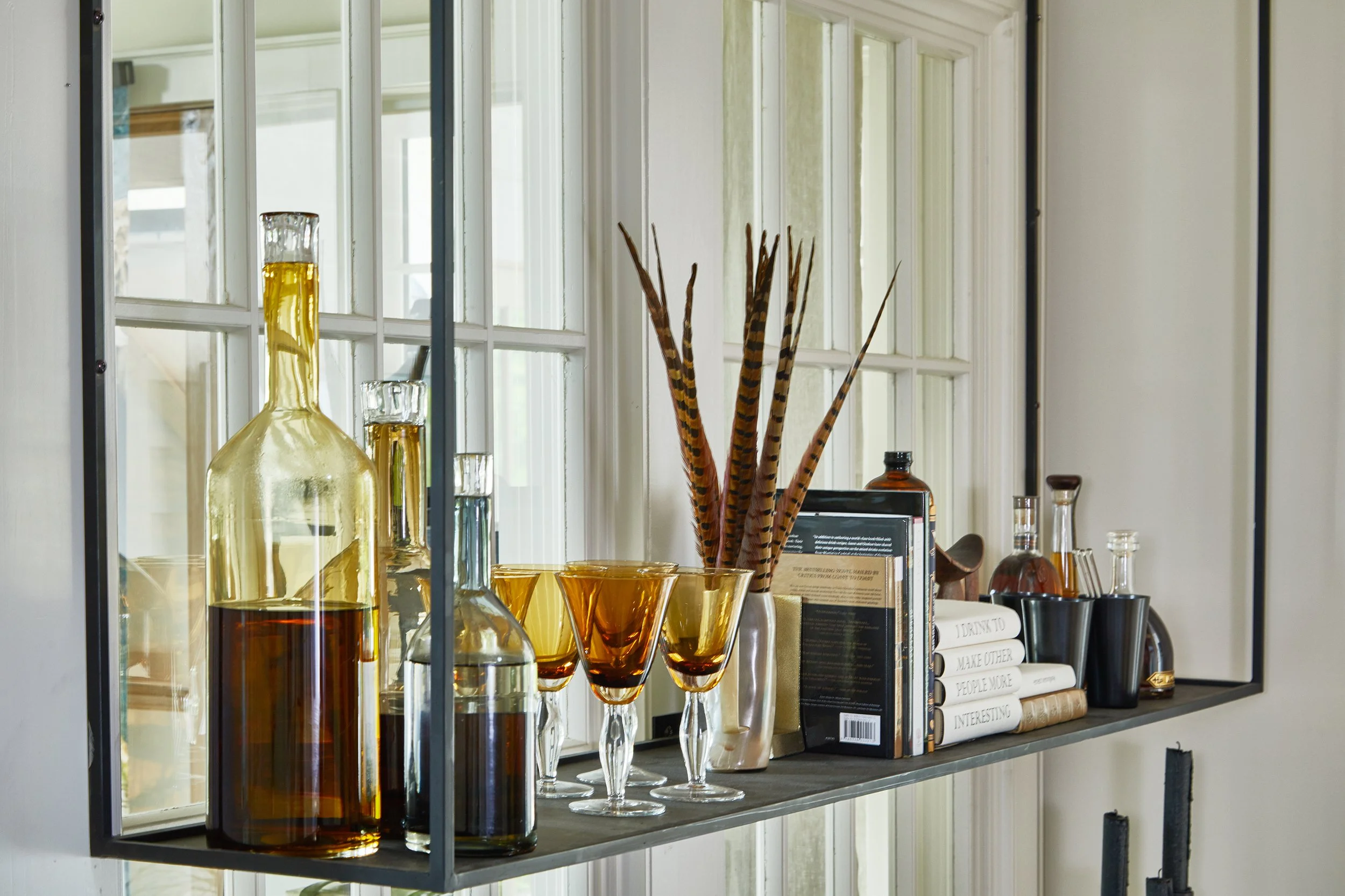 Decorative shelf with glass bottles, amber glasses, a vase with feathers, books, and small plants, placed in front of a window with white framing.