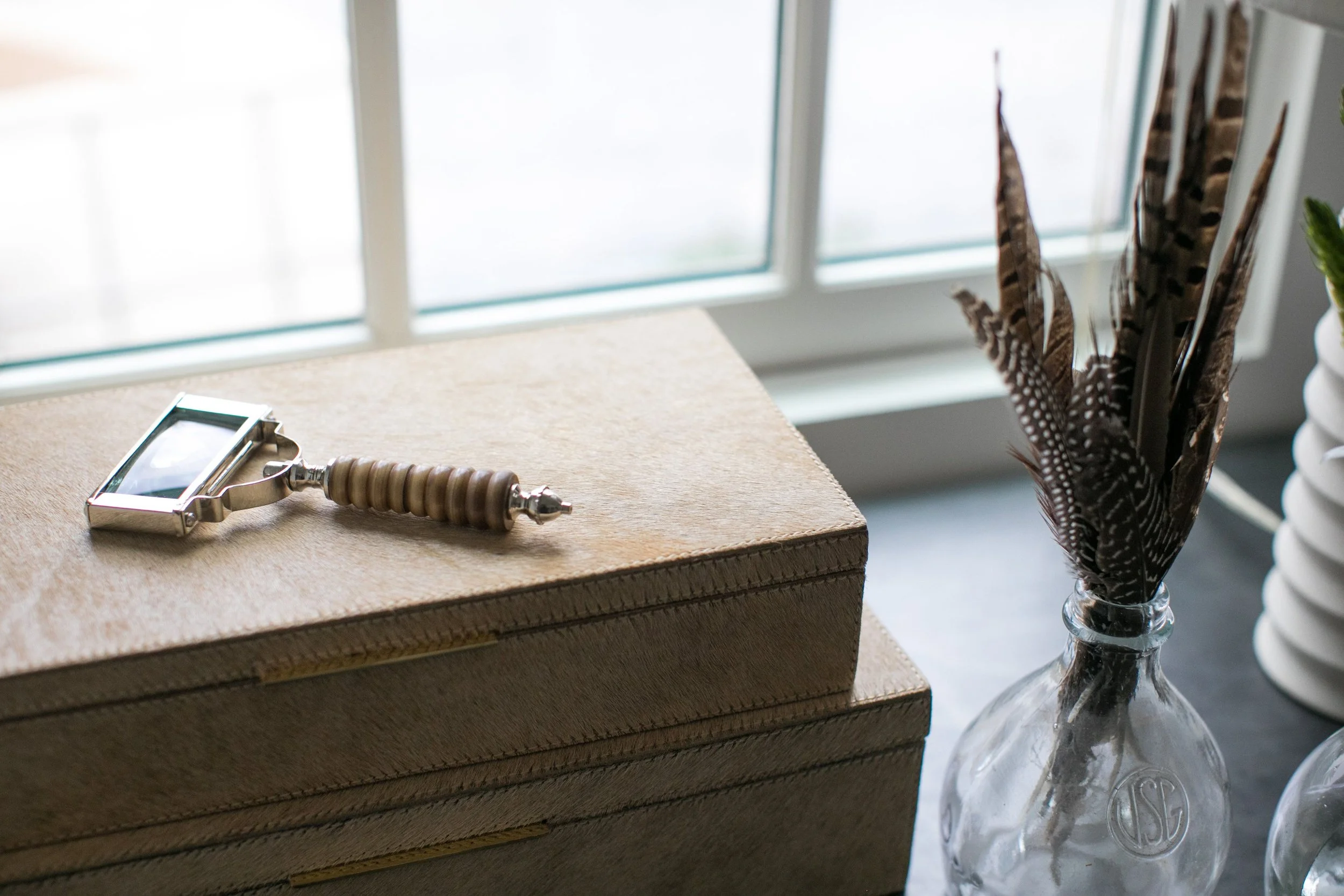 A vintage-style handheld mirror with a beaded handle rests on a closed beige textured box, beside a glass vase holding brown and white striped feathers, near a window with soft natural light.