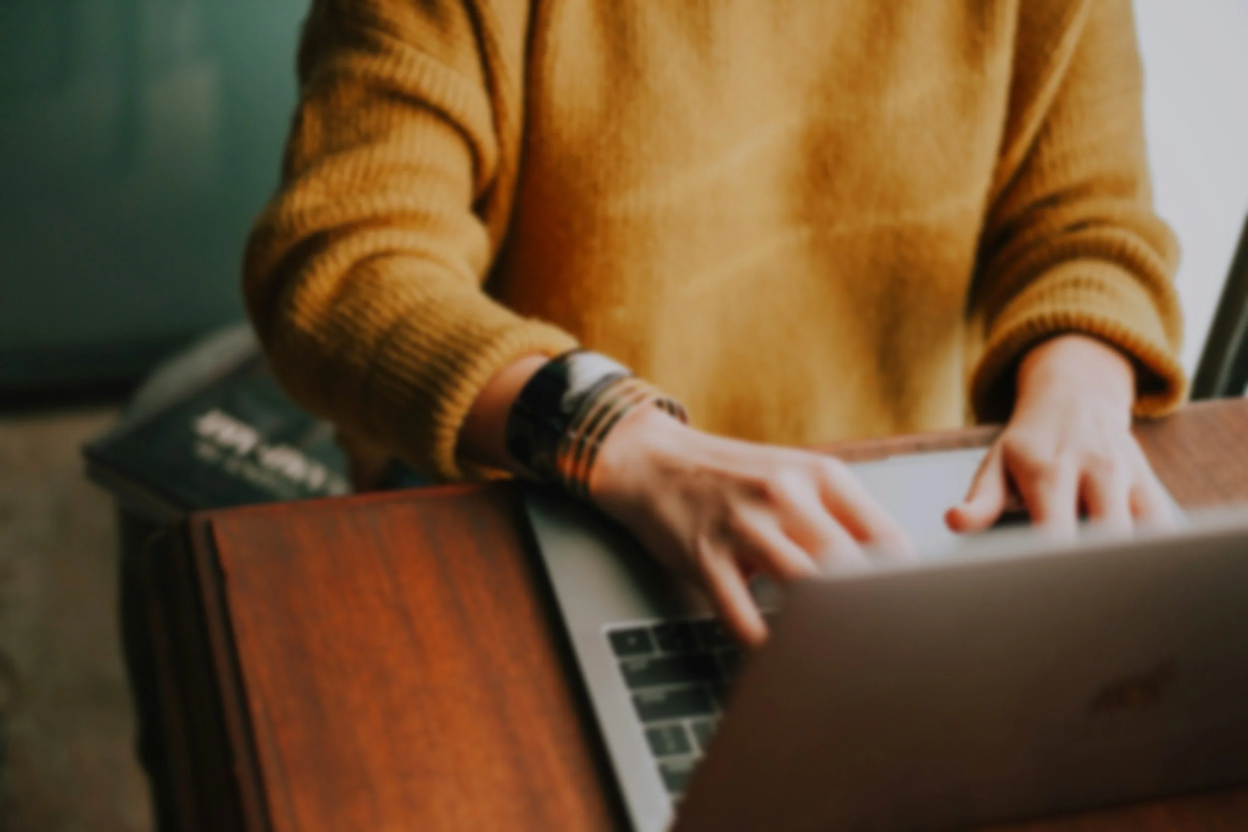Person wearing a mustard yellow sweater typing on a silver laptop, sitting at a wooden desk.