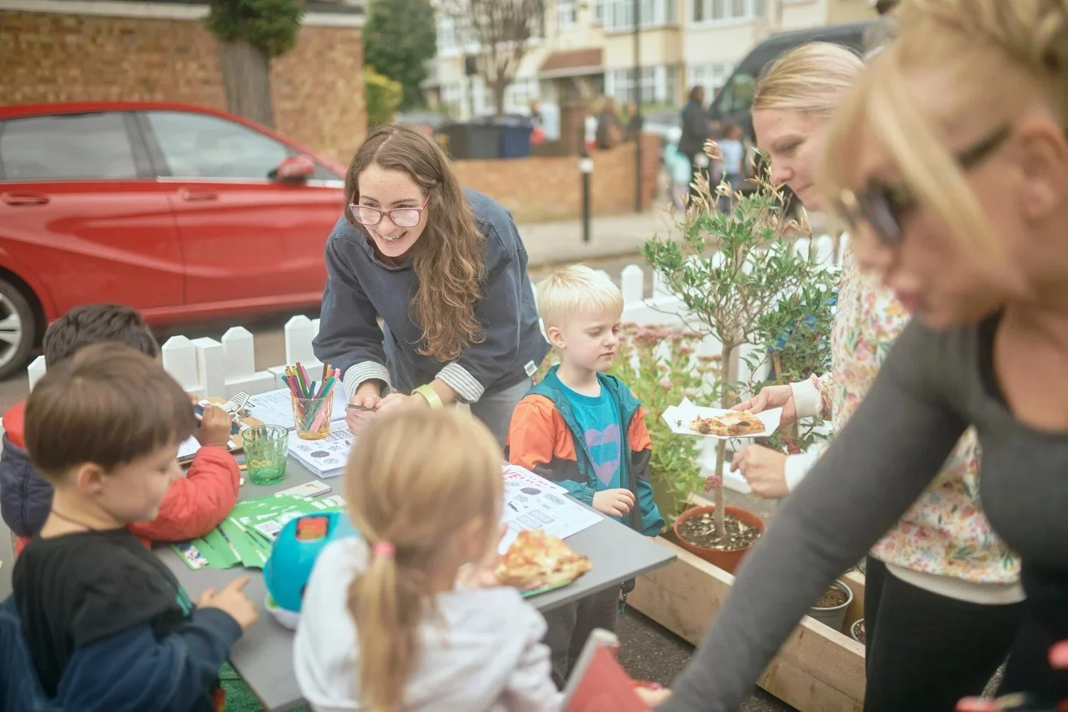 A photo of children and adults at a pop-up parklet. They are sitting at a table with pens, paper and food.