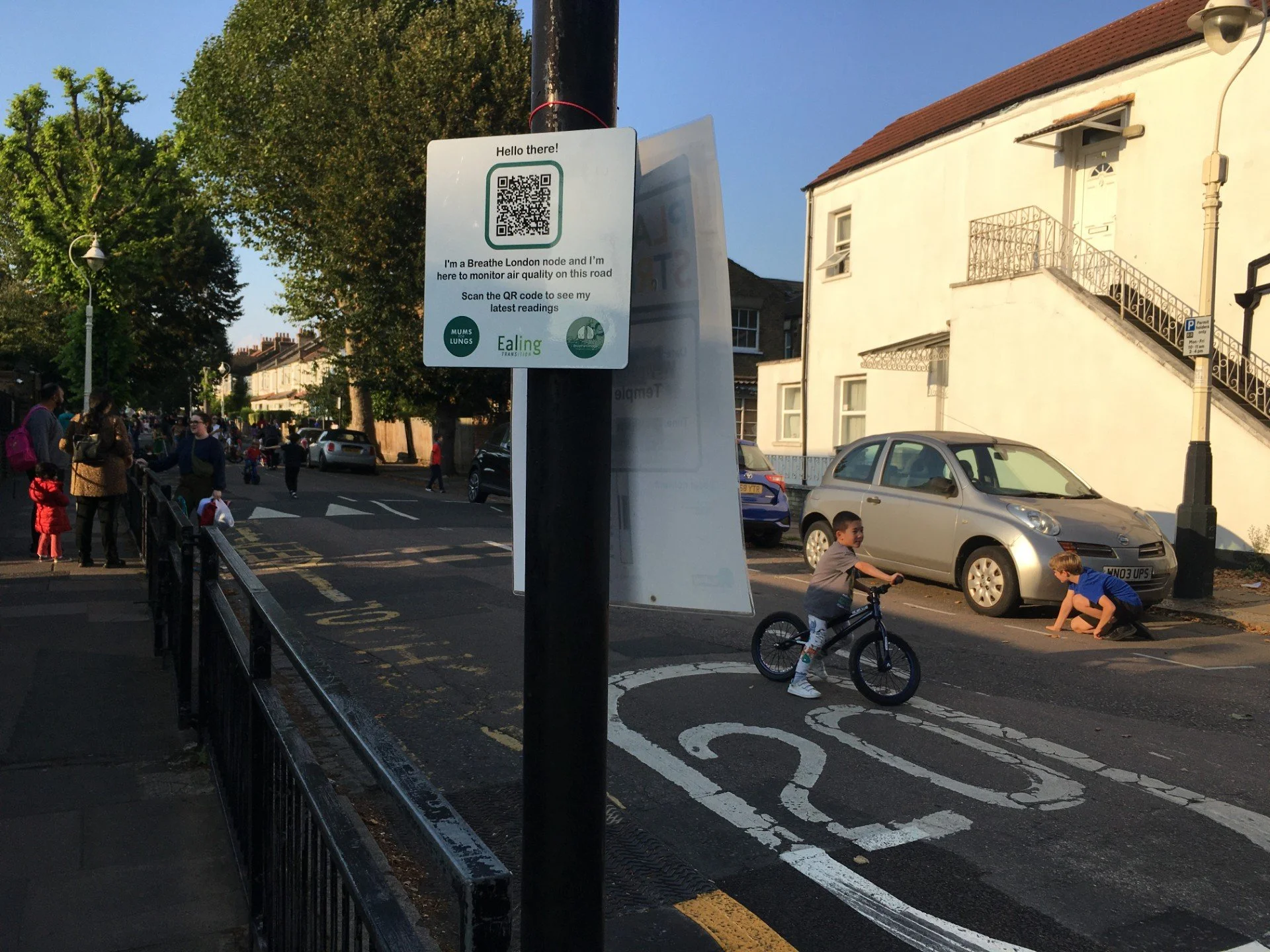 An image of a street and a sign that says "Hello there! I'm a Breathe London node and I'm here to monitor air quality on this road. Scan the QR code to see my latest readings". A child can be seen behind the sign riding a bicycle.