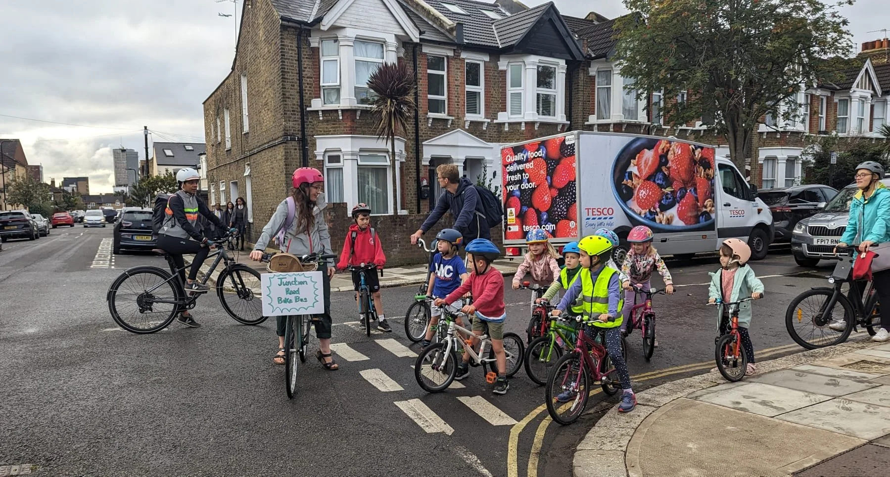 A photo of children and a few adults riding bicycles on a road. One adult's bike has a sign which reads "Junction Road Bike Bus"