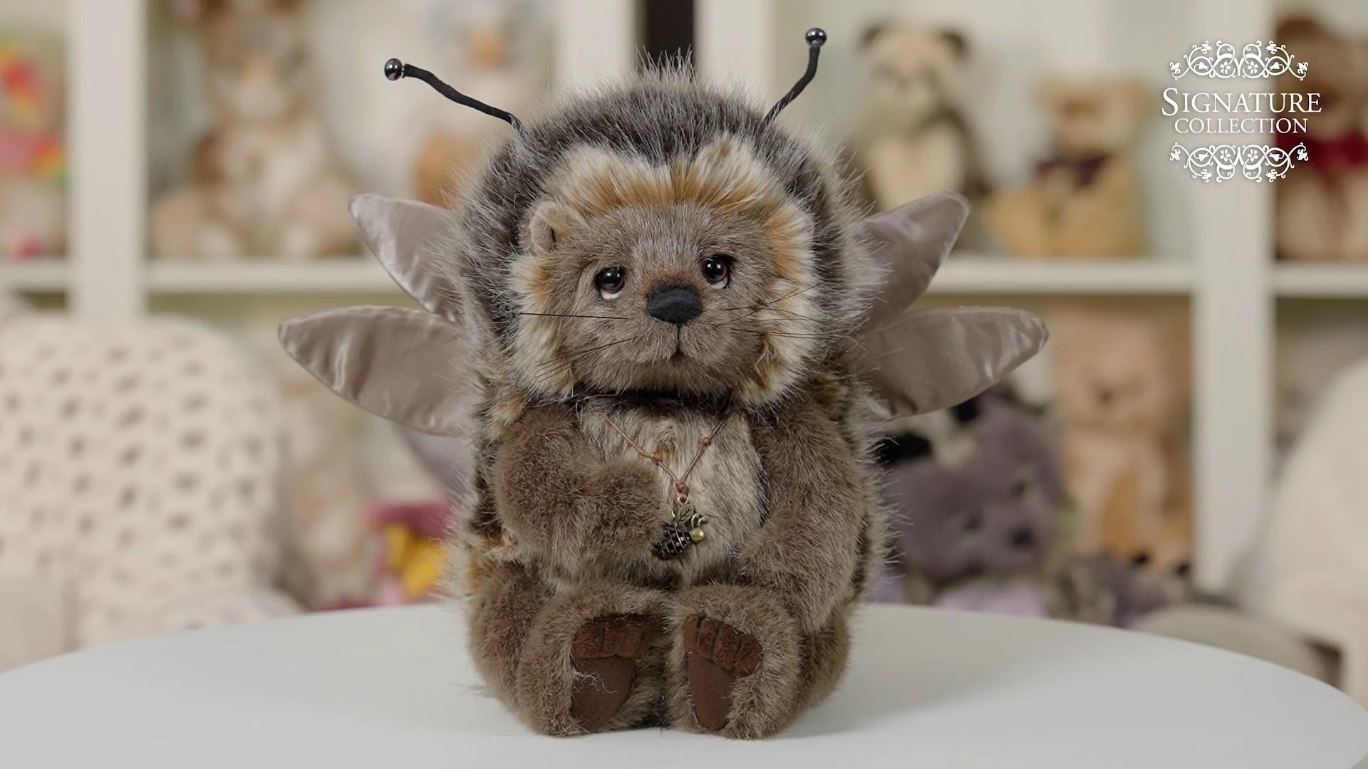 A plush toy combining a puppy and a bee, with puppy facial features, bee antennae, wings, and a necklace, sitting on a white surface with blurred stuffed animals in the background.