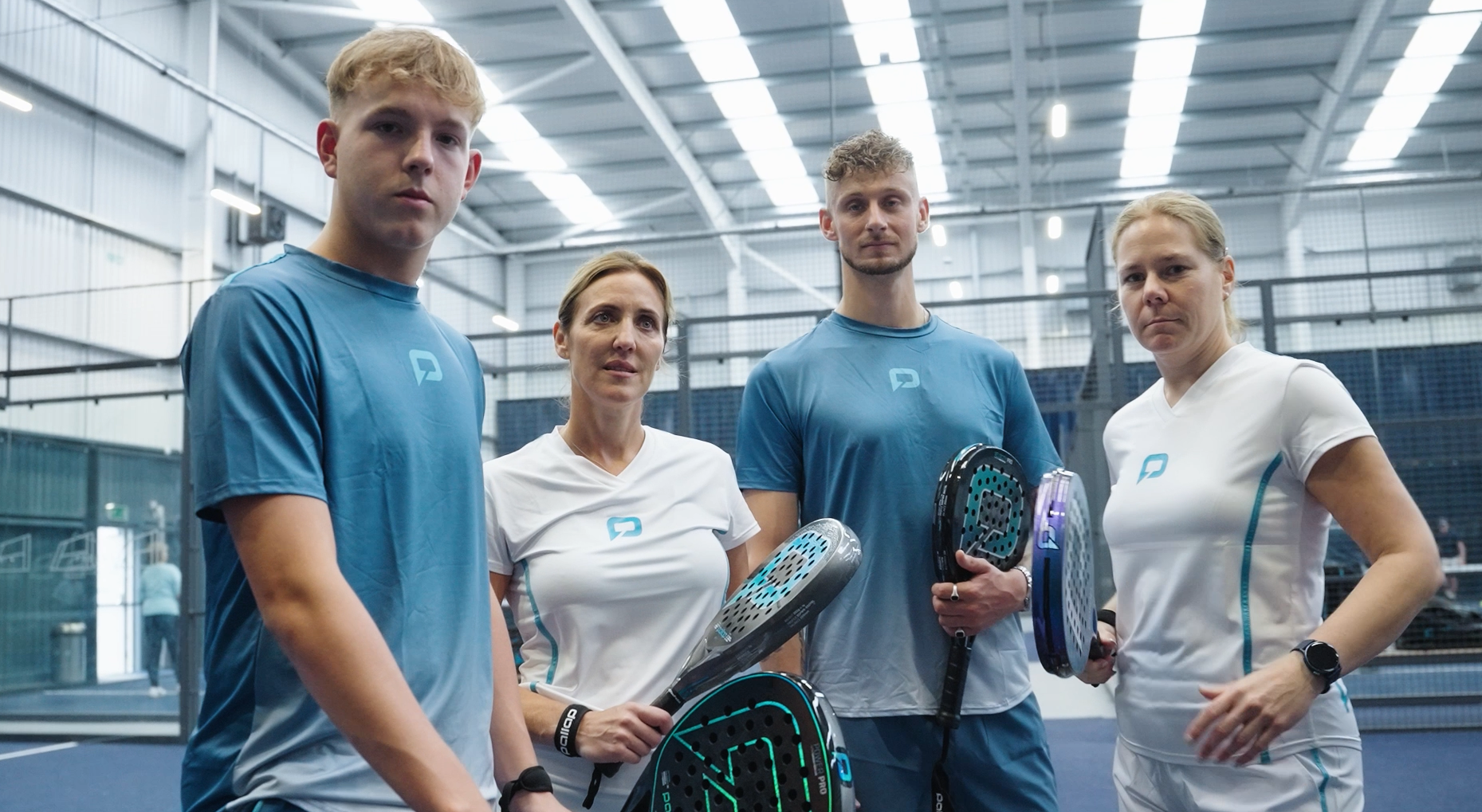 Four people standing in an indoor sports facility holding padel rackets, wearing sportswear, with a netted court background.