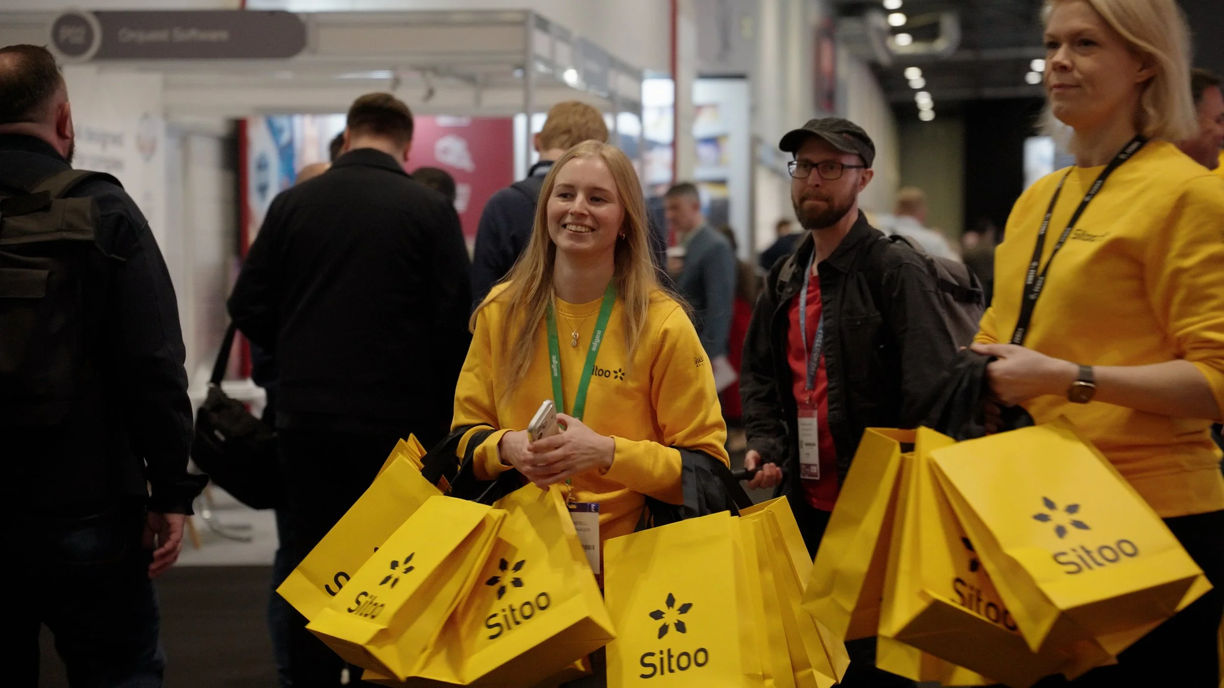 Group of people at a conference or event, standing near a booth with yellow bags labeled 'Sitoo,' engaged in conversation.