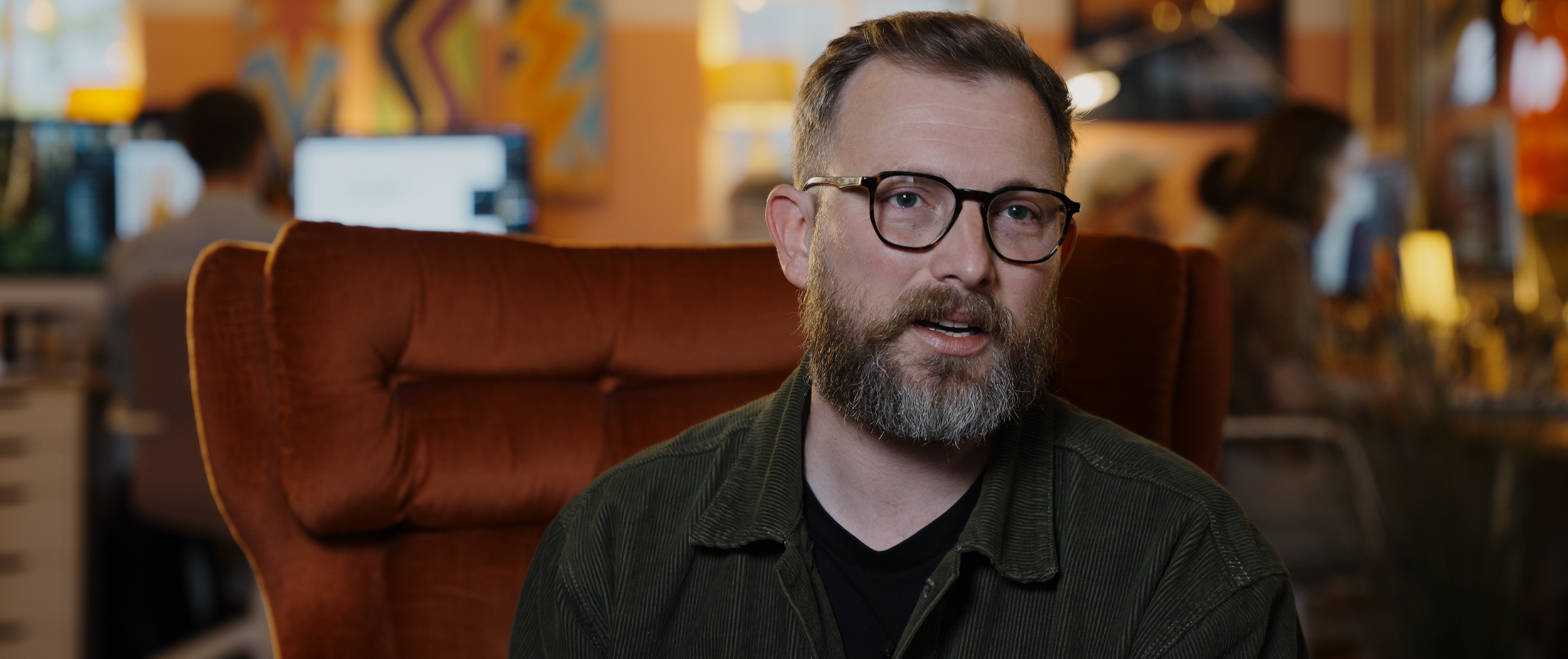 A man with glasses and a beard sitting on a chair in a colorful cafe or office space, with blurred people working on computers in the background.