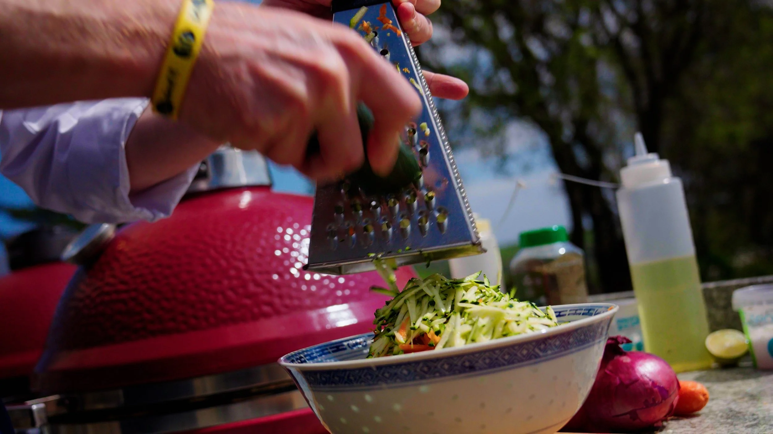 Person grating cucumber into a bowl of salad outdoors, with various bottles and food items on a table.