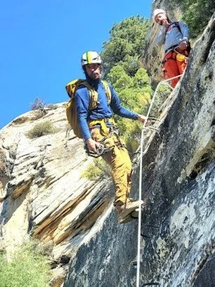Man in outdoor gear wearing a white helmet and gray shirt, smiling in a natural, rocky environment with greenery.