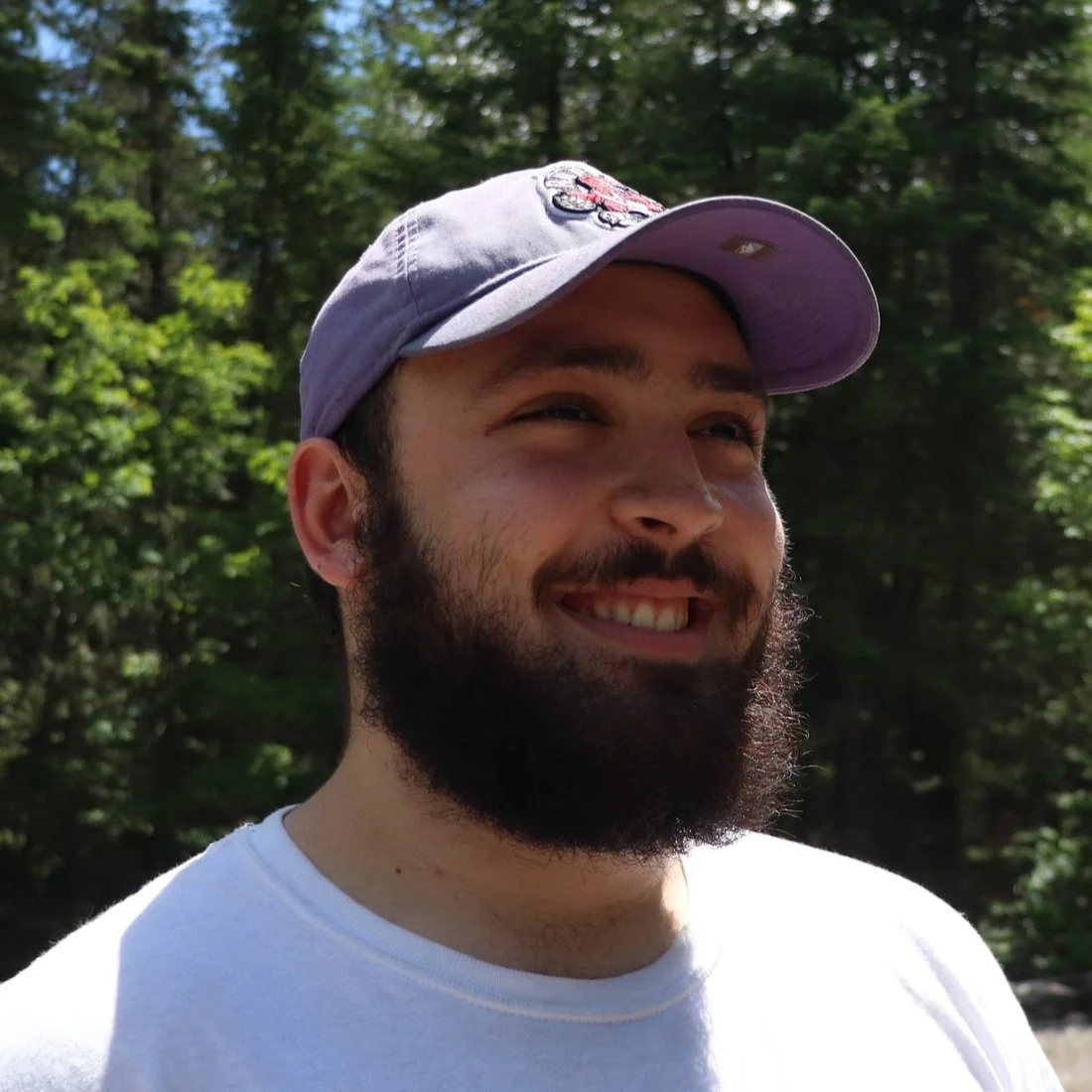 A man with a beard and mustache smiling outdoors, wearing a gray baseball cap and a white t-shirt, with a background of trees and sunlight.