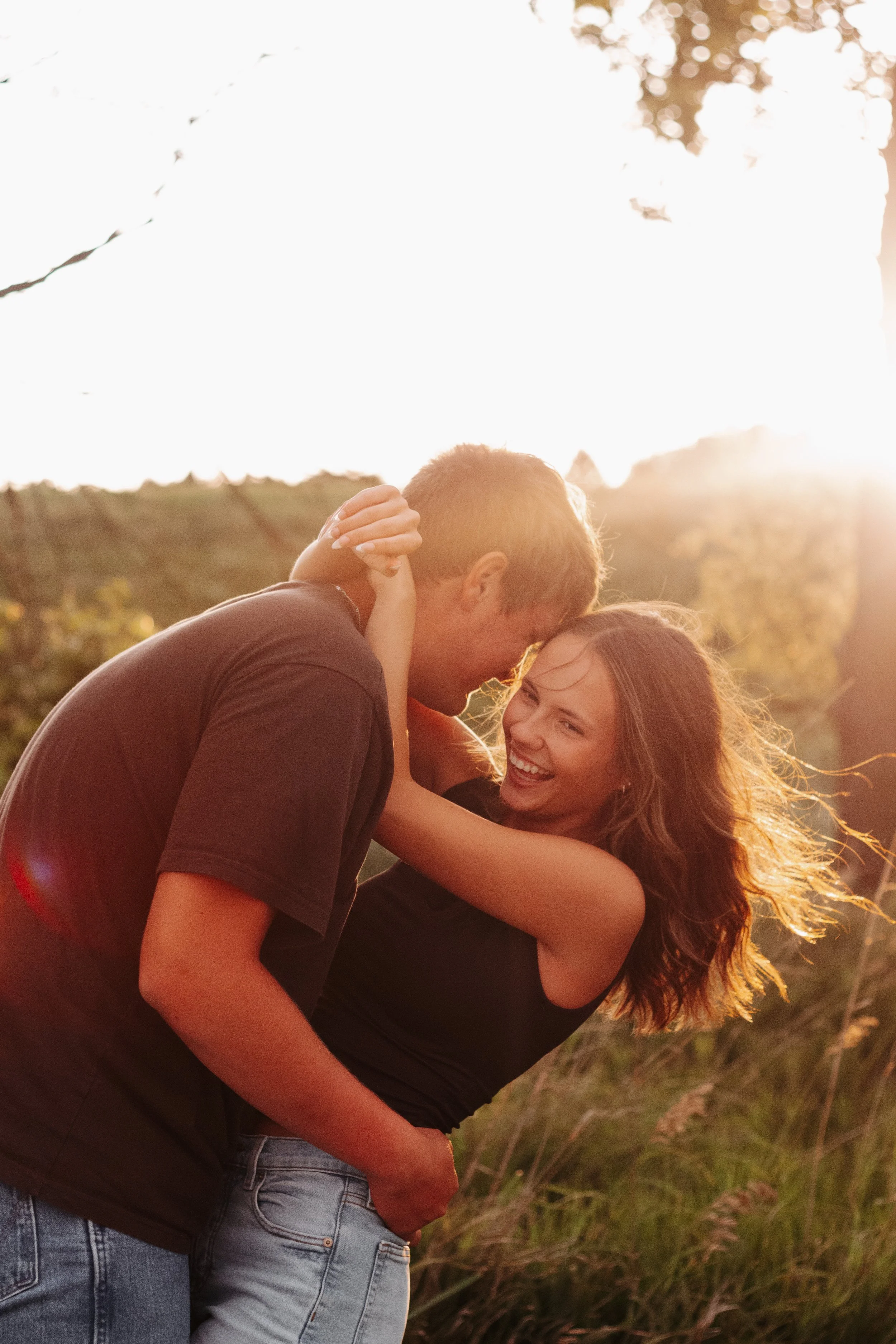 A couple happily embracing outdoors during sunset, with the man lifting the woman, both smiling.
