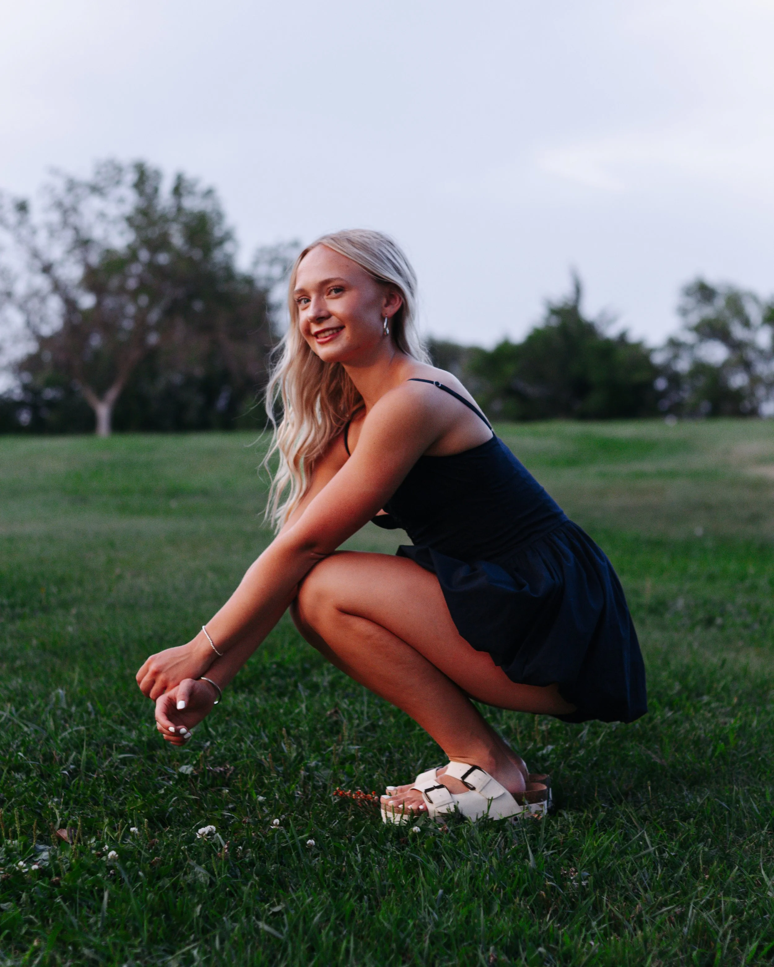 A young woman with long blonde hair wearing a navy blue dress squatting on grass in a park during sunset, smiling at the camera.
