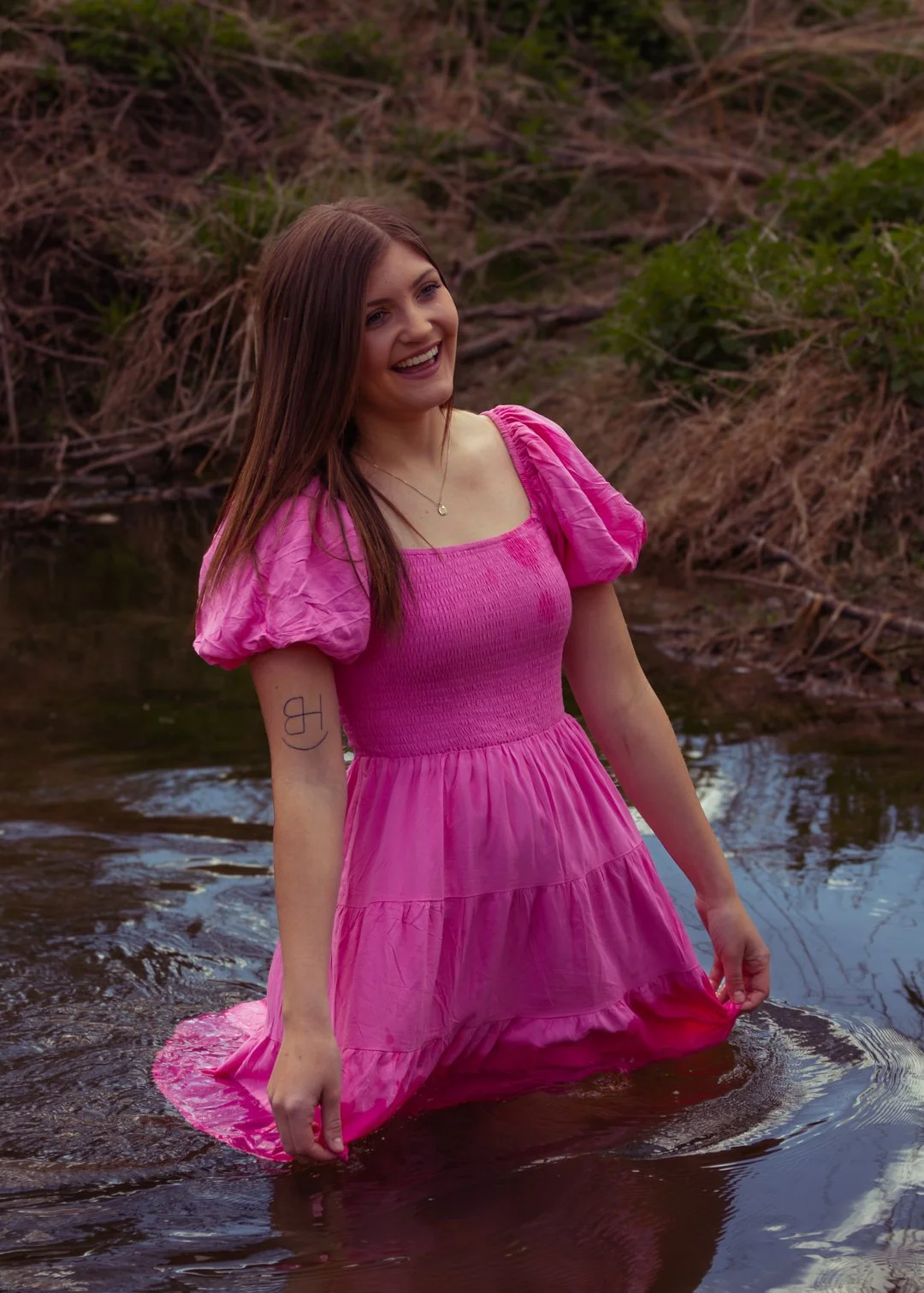 A young woman with long brown hair, wearing a bright pink dress, standing in a shallow stream with a joyful expression.