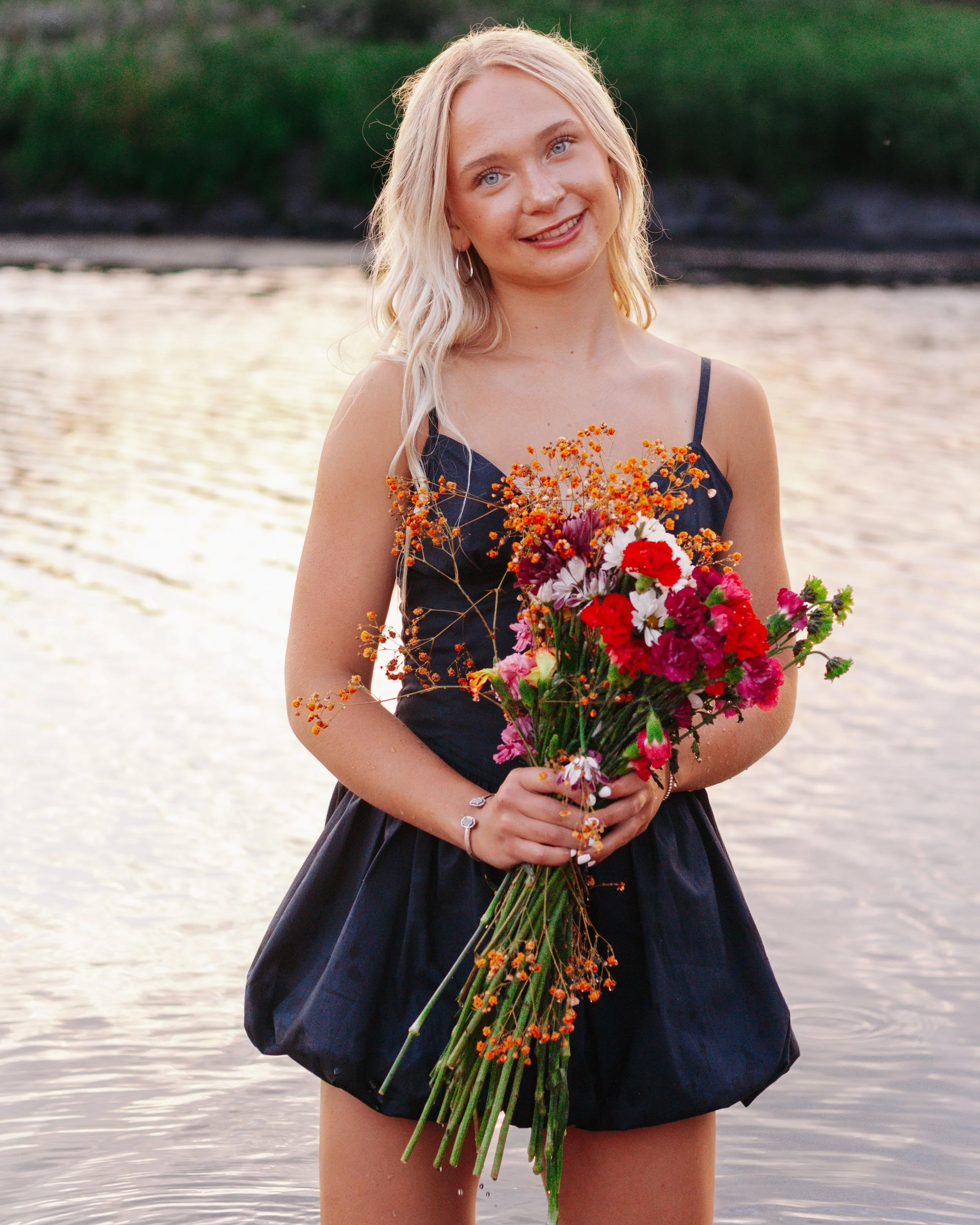 A young woman with long blonde hair wearing a black dress holding a large bouquet of colorful flowers, standing in a body of water with green trees in the background.