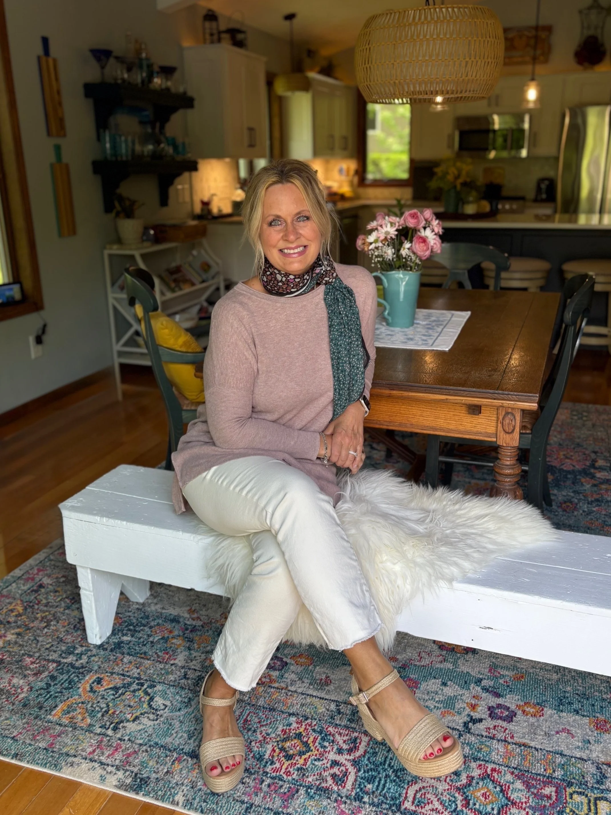 Smiling woman sitting on a white bench with a fluffy white cushion in a cozy, well-lit kitchen and dining area with flowers on the table.