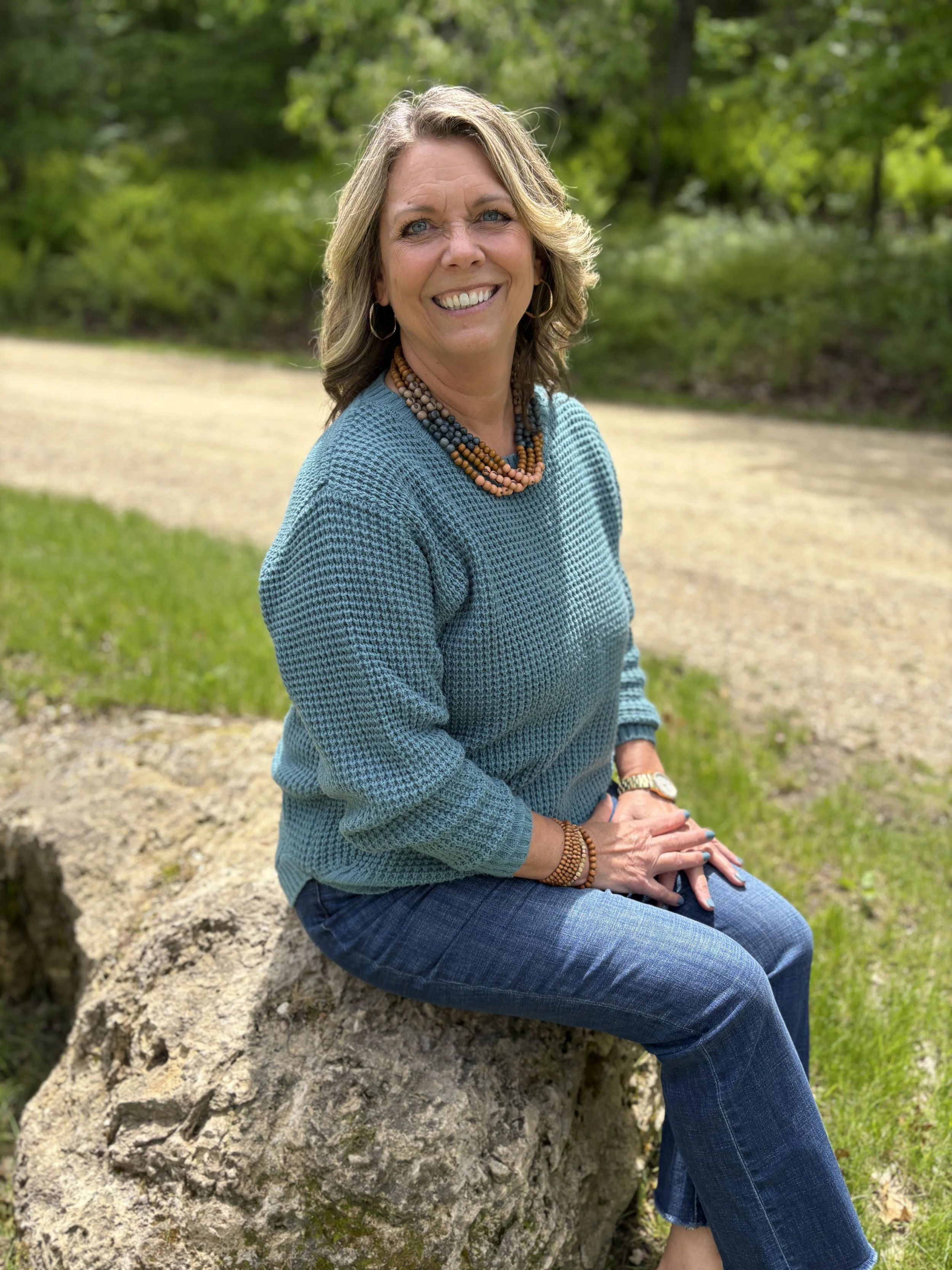A woman with shoulder-length light brown hair, wearing a turquoise sweater and blue jeans, sitting on a large rock outdoors with green trees in the background. She is smiling and wearing beaded necklaces, bracelets, a watch, and hoop earrings.