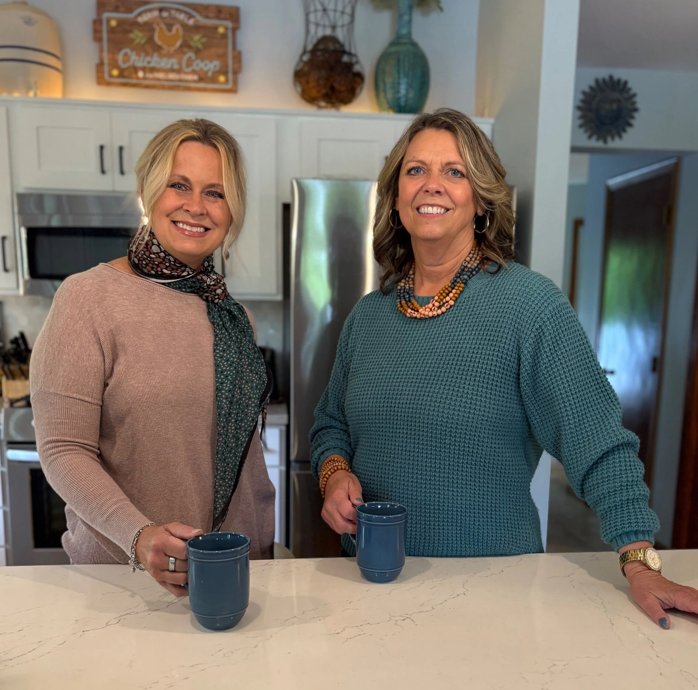 Two women smiling and holding blue mugs in a kitchen. One woman has blonde hair and a patterned scarf, the other has gray hair and is wearing colorful necklaces.