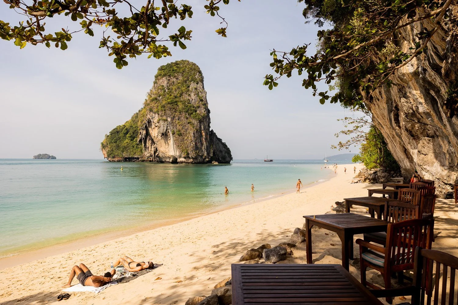 A tropical beach scene with a large limestone island in the ocean, people swimming, relaxing on the sand, and shaded wooden tables and chairs under trees on the shoreline, in Thailand.