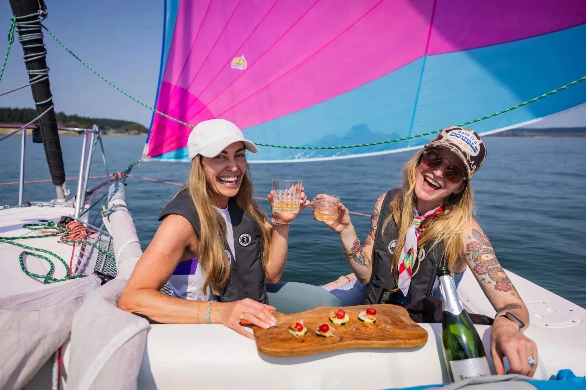 A sailboat named 'WINDY' anchored near the rocky shore with a swimmer approaching from the water during daylight, with a forested island and cloudy sky in the background in Penn Cove.