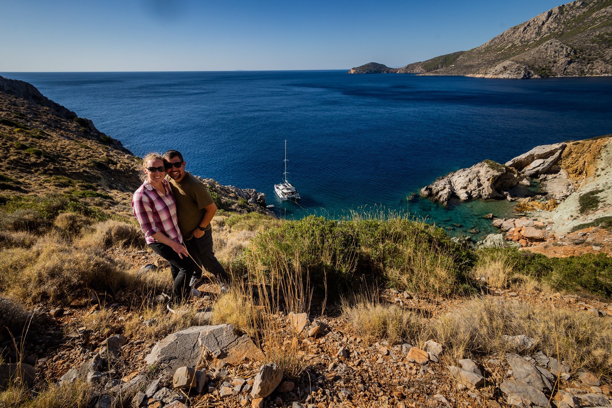 A smiling couple standing on a rocky hillside overlooking a deep blue ocean with a sailboat anchored near rocky cliffs, with hills in the background on a clear sunny day.