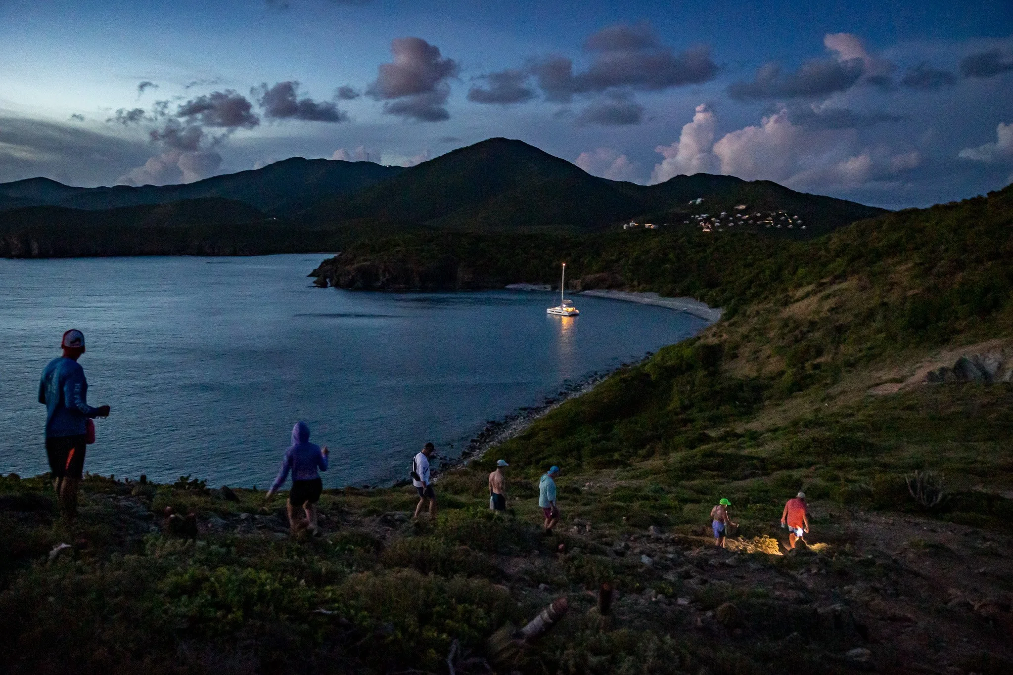 Group of hikers walking along a rocky trail near a body of water at dusk, with mountains and a boat anchored in the water in the background.
