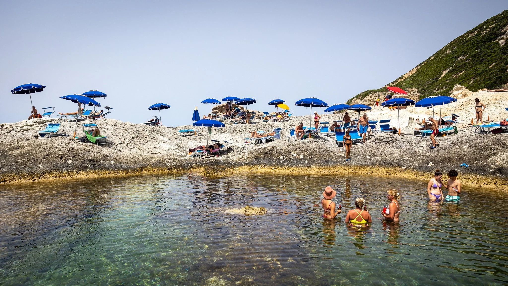 People relaxing on rocky beach with blue umbrellas, some swimming and standing in the clear water, and others sunbathing on lounge chairs under umbrellas on the Island of Ponza.