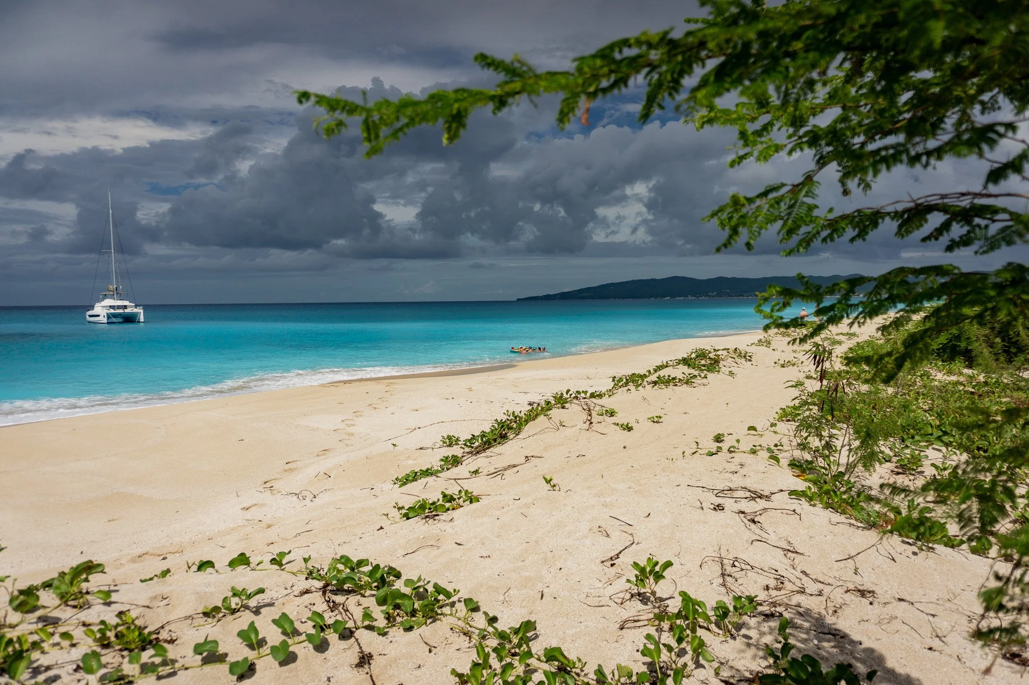 A beach scene with white sand, turquoise water, a sailboat anchored near the shore, and an island in the distance. Cloudy sky with overcast weather and green foliage partially framing the image.