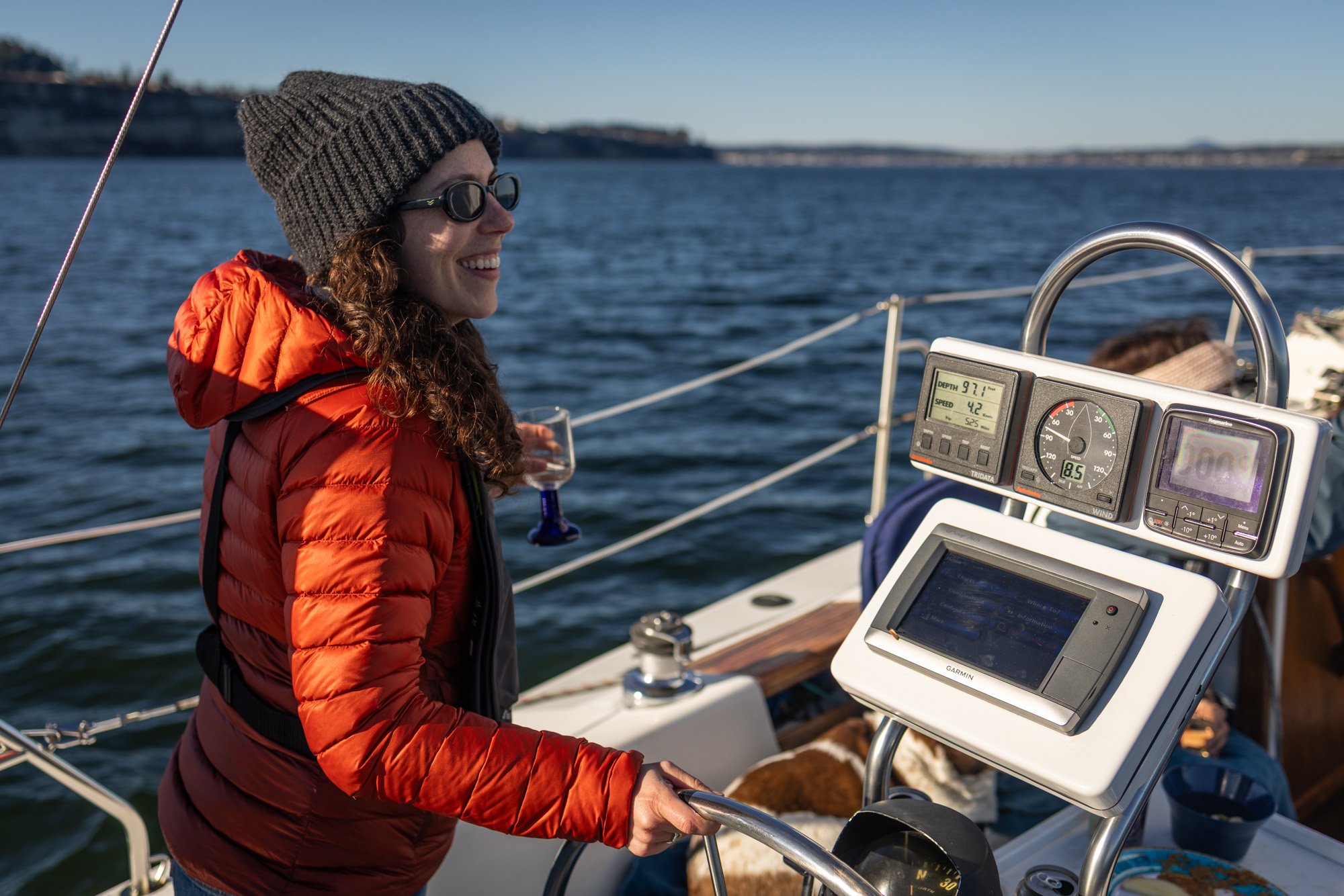 A sailboat named 'WINDY' anchored near the rocky shore with a swimmer approaching from the water during daylight, with a forested island and cloudy sky in the background in Penn Cove.
