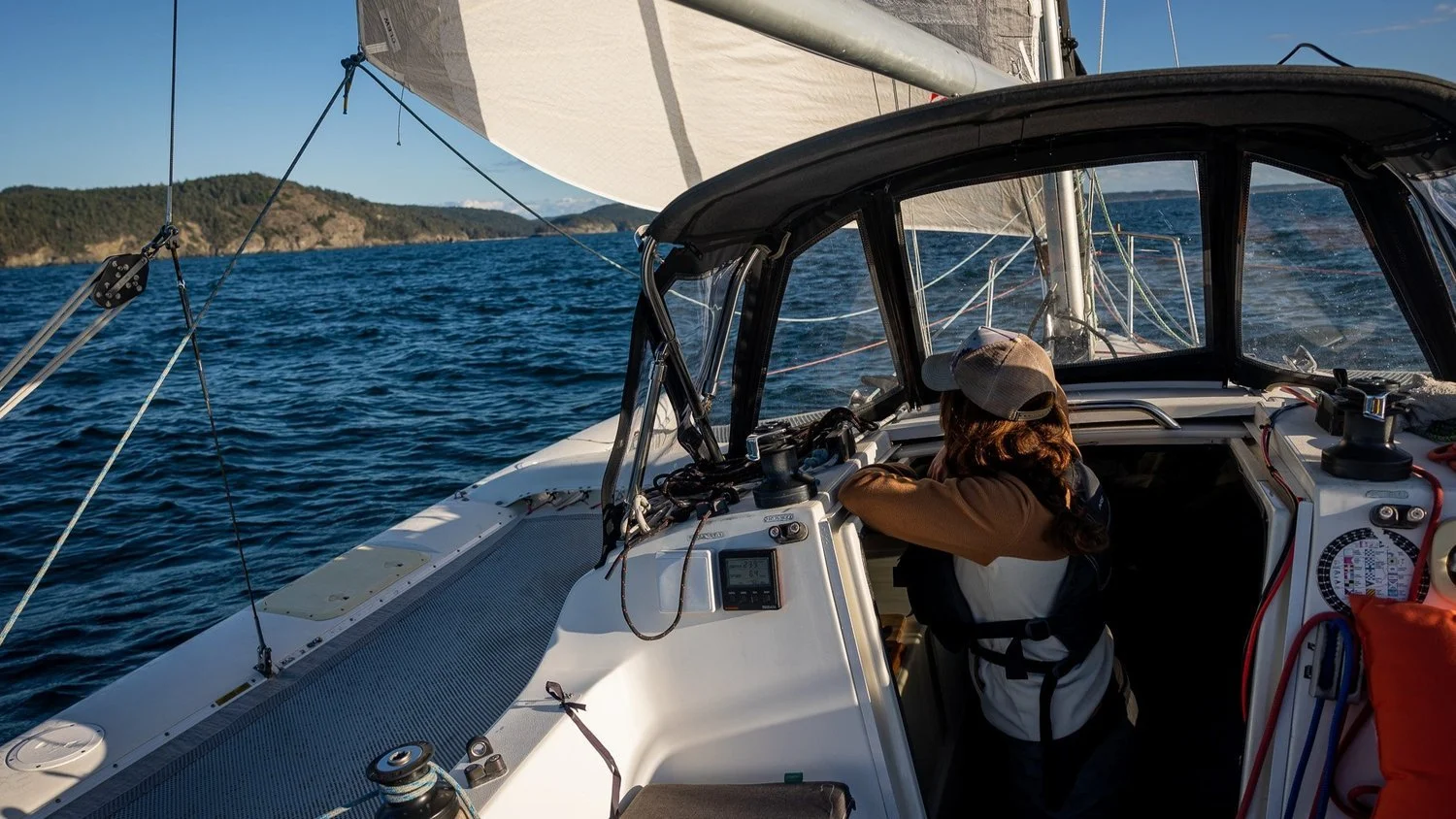 Person sailing a boat on open water with land visible in the background.
