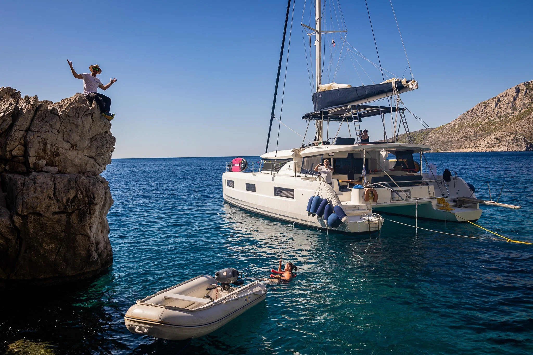 A person sitting on a rocky cliff next to a boat and a large sailboat anchored in the water, with other people swimming nearby and mountains in the background.