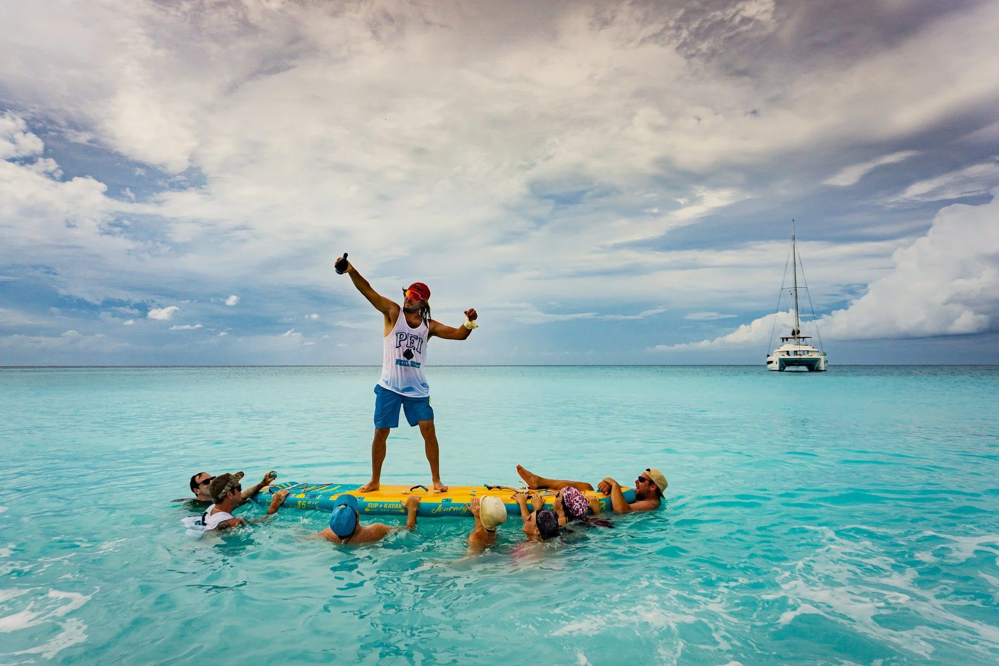 A group of people on a paddleboard in clear blue ocean water, with one man standing and holding a drink in the air, and several others around him holding onto the paddleboard. There is a sailboat in the background under a cloudy sky.