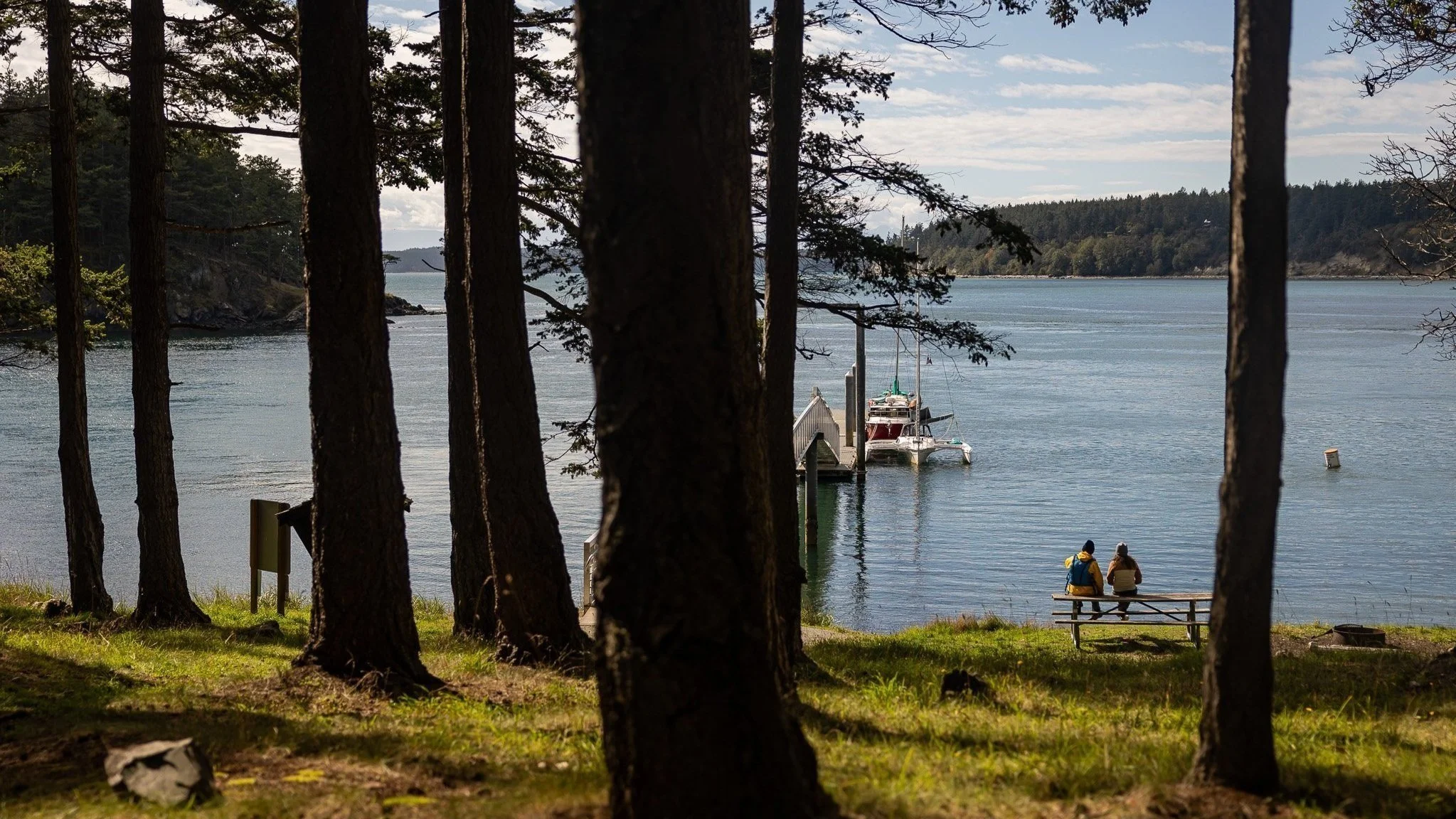 Two people are sitting on a bench near the water, on James Island in the San Juan Islands, with trees and boats in the background on a calm day.