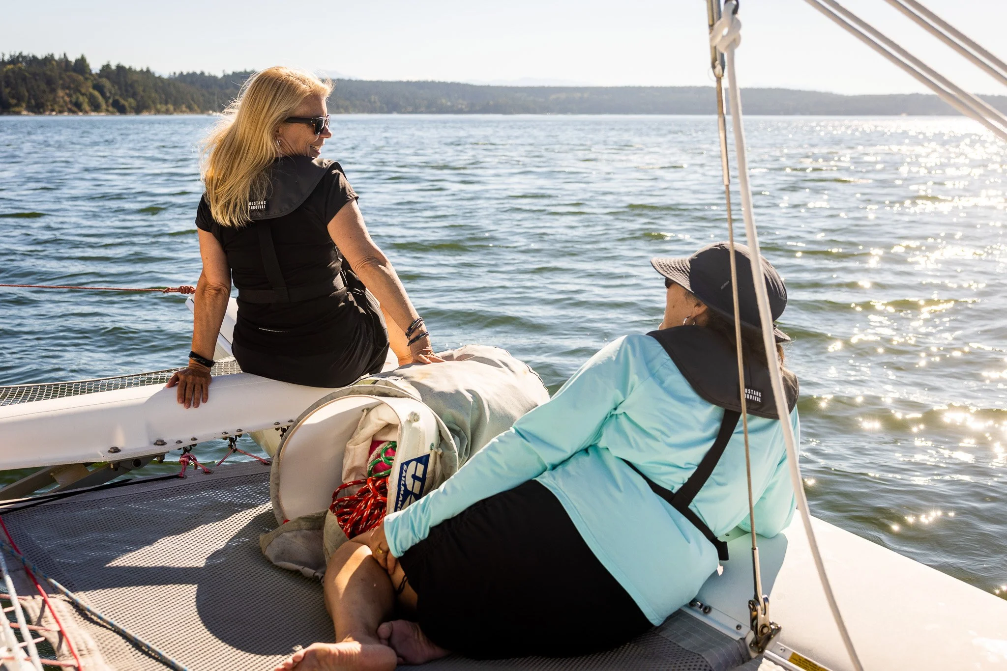 Two women sitting on a boat deck, relaxing by the water with a distant shoreline and trees, enjoying a sunny day In Penn Cove. 