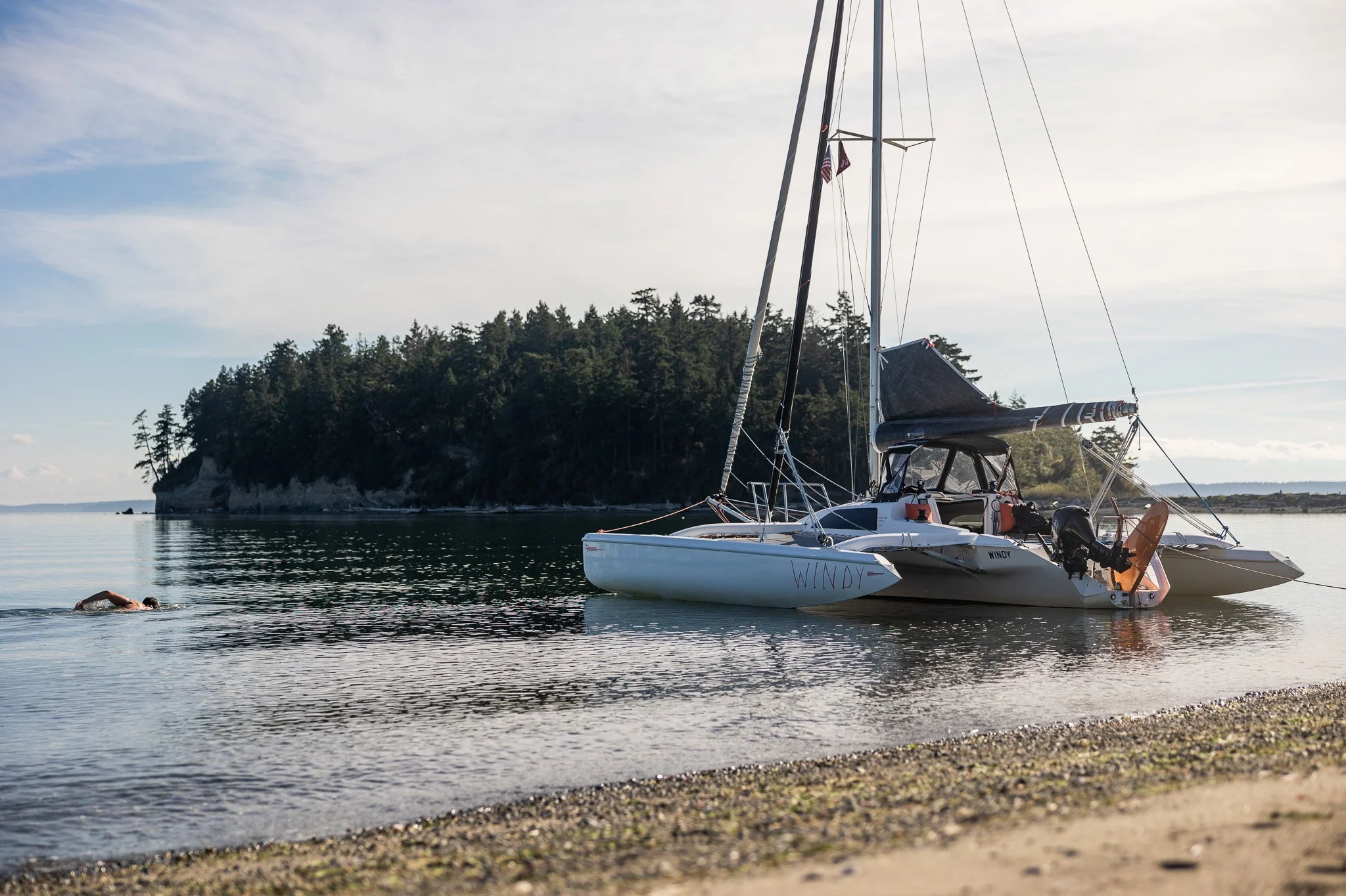 A sailboat named 'WINDY' anchored near the rocky shore with a swimmer approaching from the water during daylight, with a forested island and cloudy sky in the background in Penn Cove.