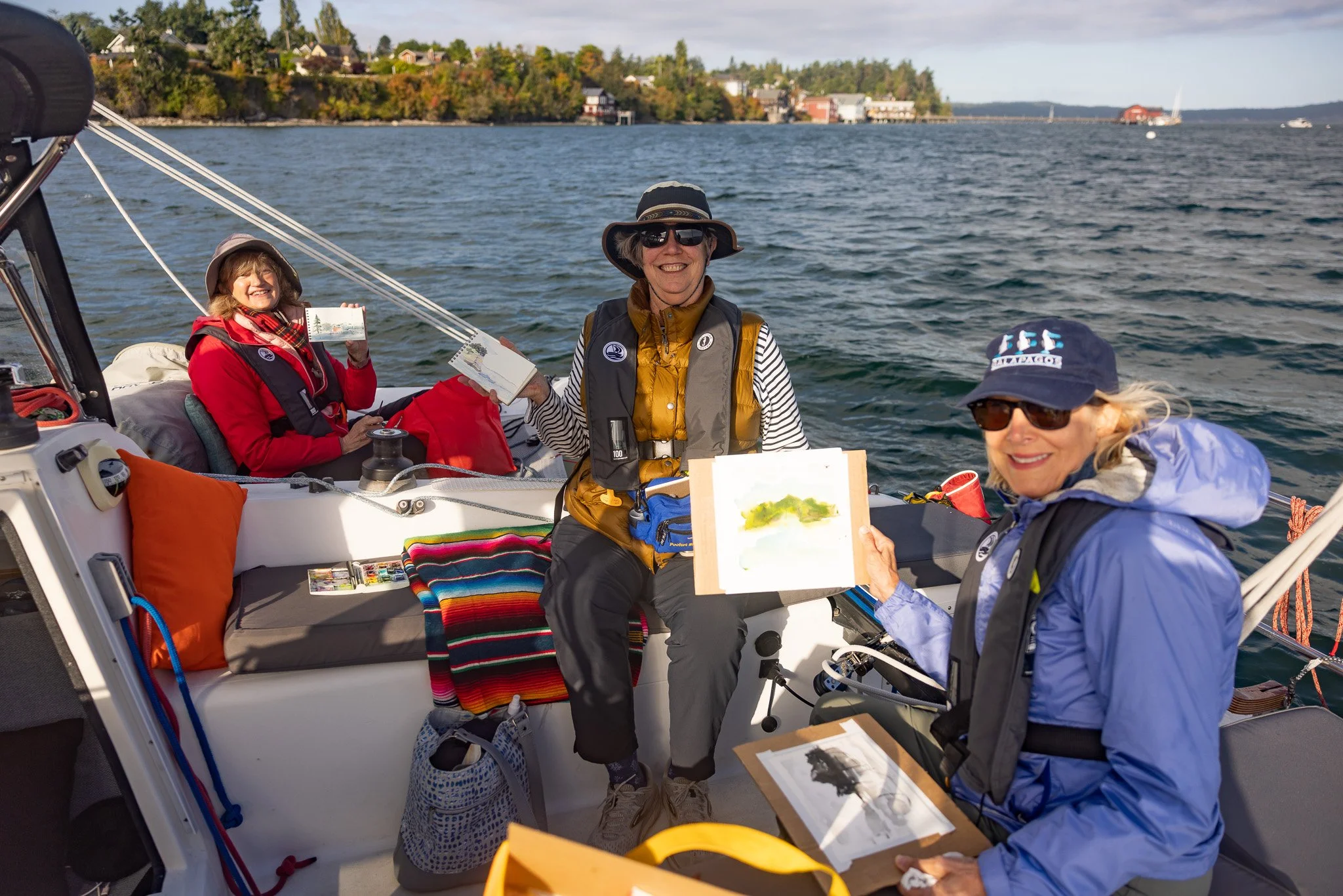 Three women on a boat, smiling and holding watercolor paintings they created, with water and trees in the background.