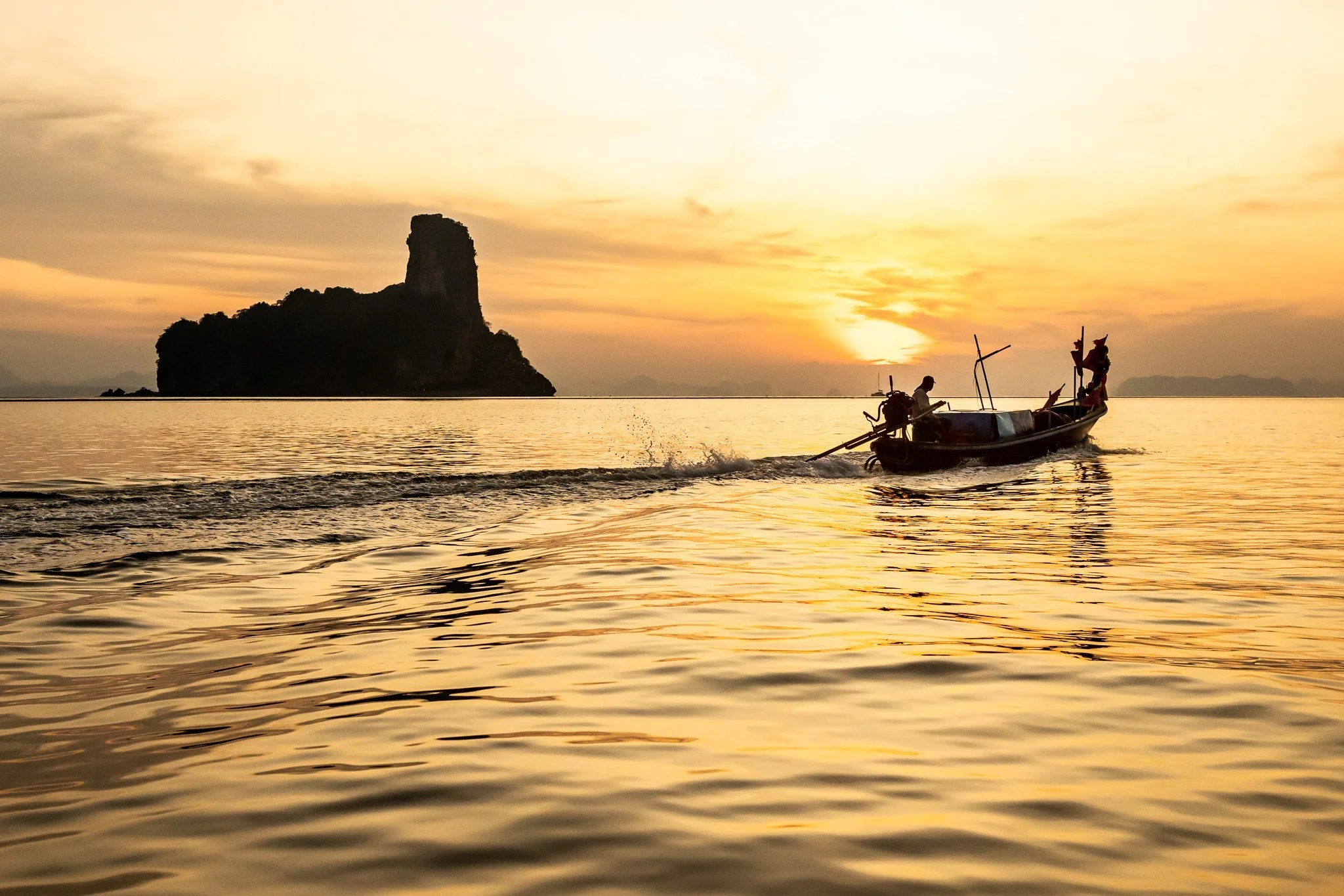 A boat with two people on the water at sunset, with a large rock formation in the background and sky filled with warm colors.