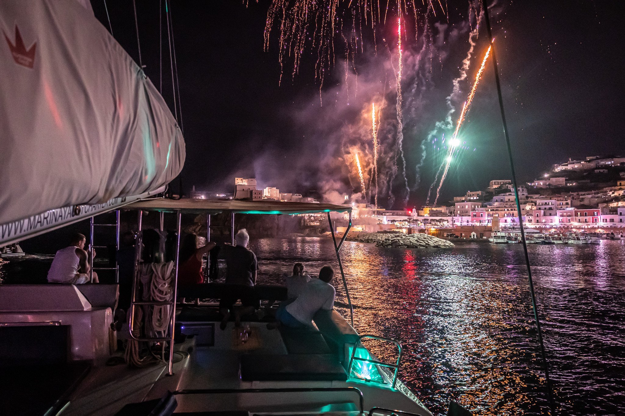 People on a boat watching fireworks over a coastal town at night.
