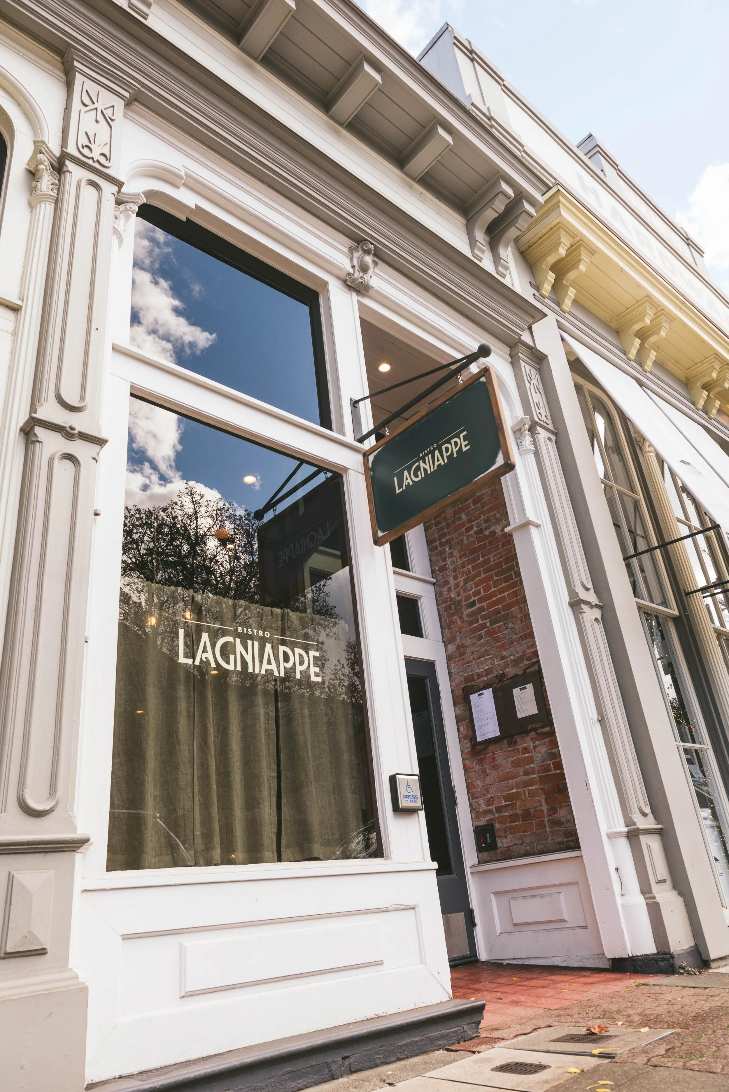 Exterior of Bistro Lagniappe with white decorative trim, large window with curtains, a signboard, and a brick wall section.