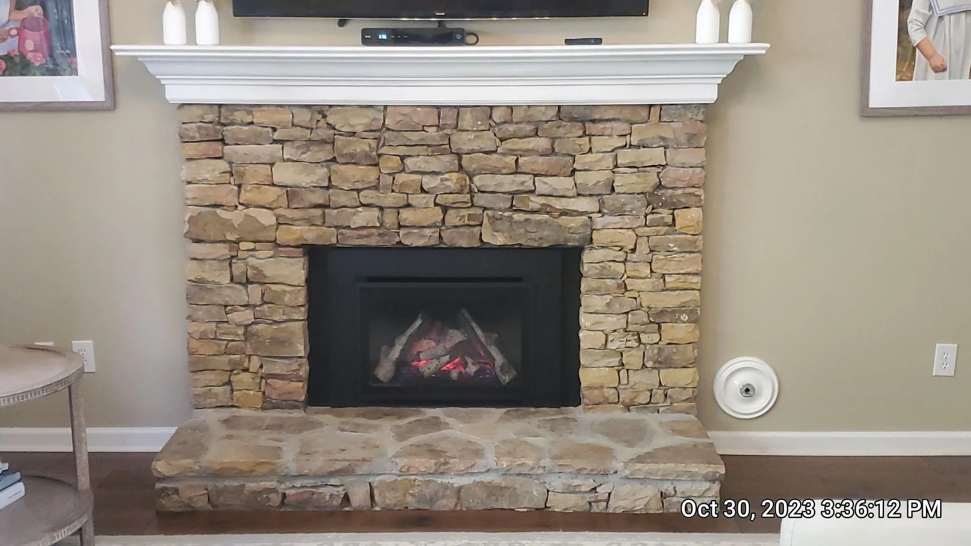A living room fireplace with a stone surround and a white mantle. Two white vases sit on the mantle, along with a black device and remote control. To the left, part of a side table is visible, and on the right side, a framed picture. The fireplace has a built-in gas logs with a glowing ember effect. The date and time on the image are October 30, 2023, at 3:36 PM.