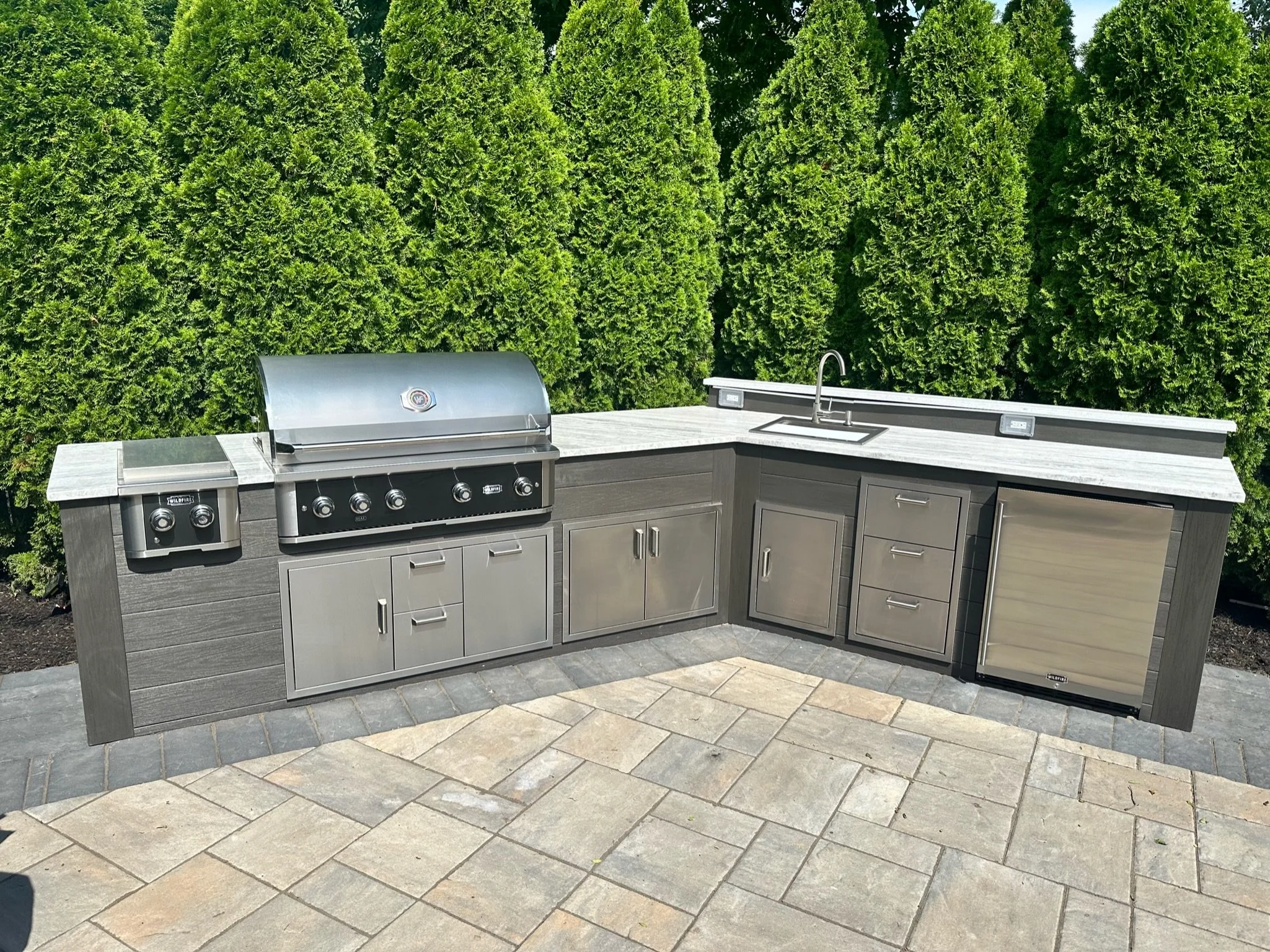 Outdoor kitchen with a grill, cabinets, a sink, and a mini refrigerator, set against a backdrop of tall green bushes.