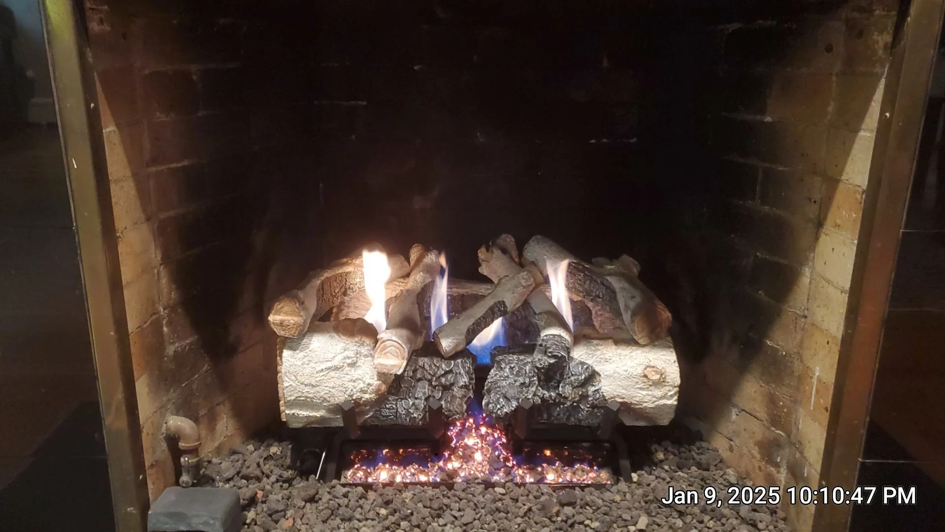 A lit wood-burning fireplace with bricks on the back and sides, filled with burning logs and glowing embers at the bottom. The fire produces bright flames and a cozy ambiance.