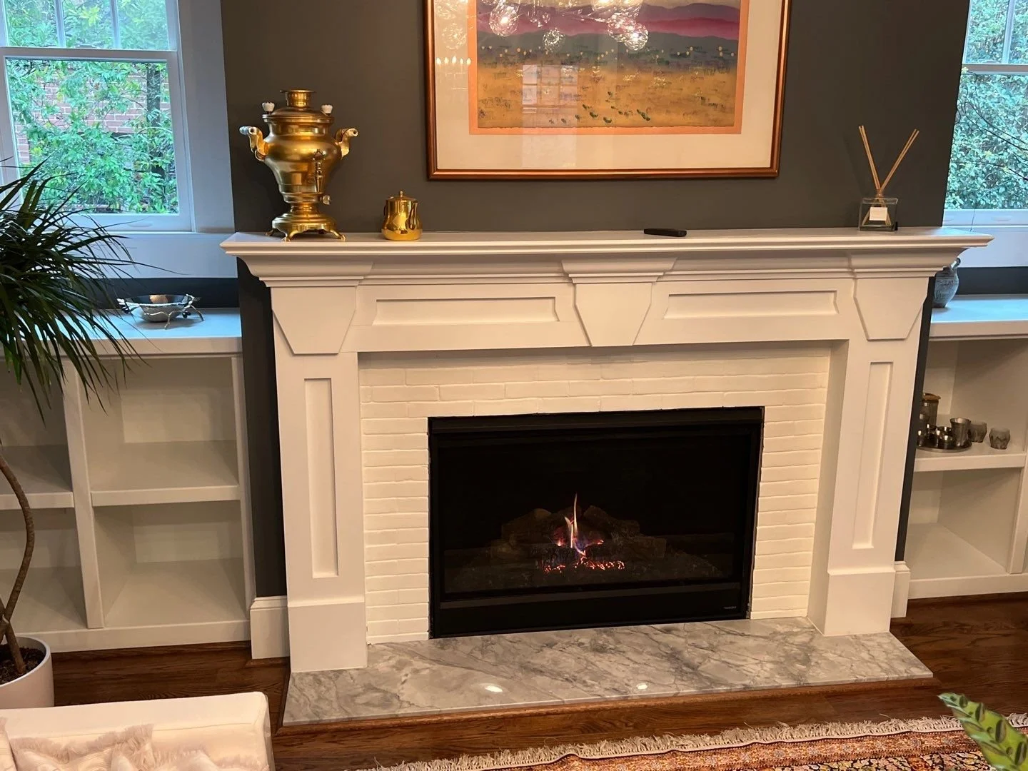 Living room fireplace with white mantel, framed artwork above, and decorative items on the mantel. Two windows flank the fireplace, showing green foliage outside. The floor is wooden with a marble hearth.
