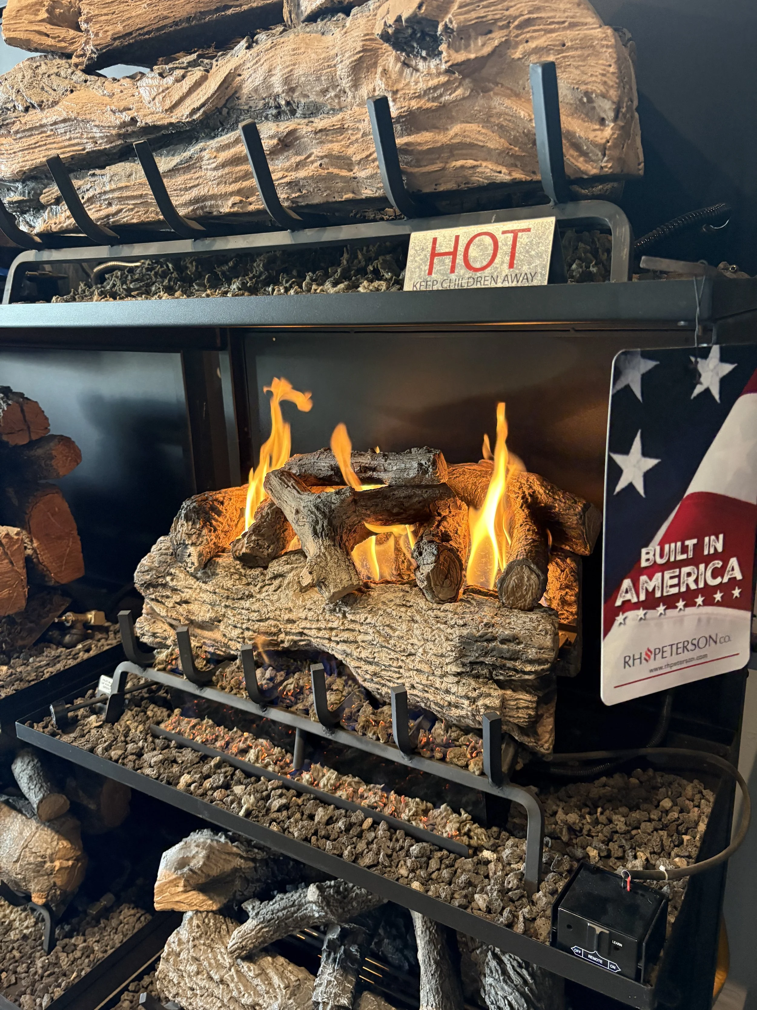 A display of artificial logs with a realistic fire, promotional signs including one labeled 'HOT KEEP CHILDREN AWAY' and another with an American flag design reading 'BUILT IN AMERICA,' and a sign displaying 'R H PETERSON Co.'